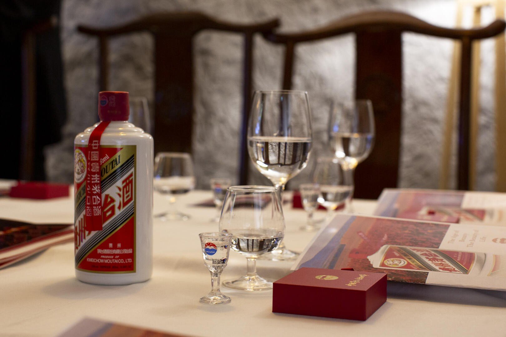 Ein weißer Tisch mit einer Flasche Moutai-Schnaps, mehreren Wassergläsern, einem Marken-Schnapsglas, roten Geschenkboxen und Broschüren vor Holzstühlen und einer strukturierten Wand.