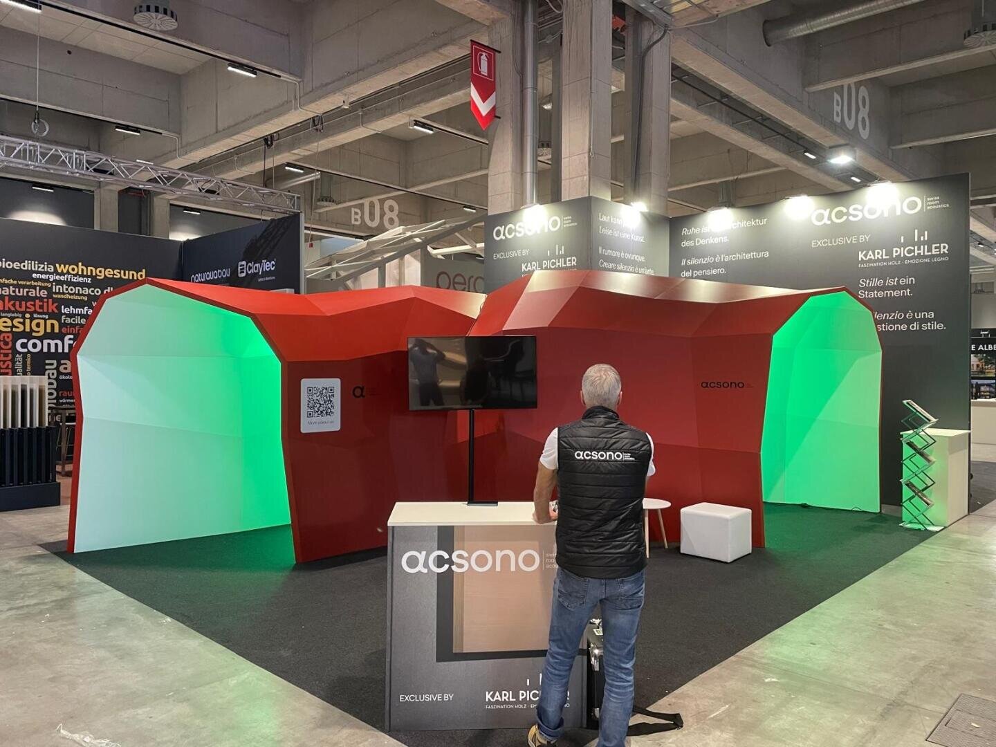 Ein Mann steht vor einem modernen Messestand mit rot und grün beleuchteten Wänden, einem Fernsehbildschirm und einem Acsono-Schild auf einer Fachmesse. Der Raum hat hohe Decken und andere Stände im Hintergrund.