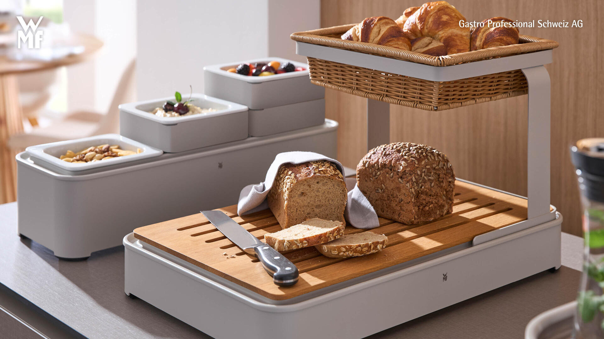 Un buffet de petit-déjeuner comprenant une miche de pain et des tranches sur une planche à découper avec un couteau, une corbeille de croissants et trois plateaux avec des fruits, des yaourts et des noix. Tous les articles sont exposés dans une cuisine moderne.