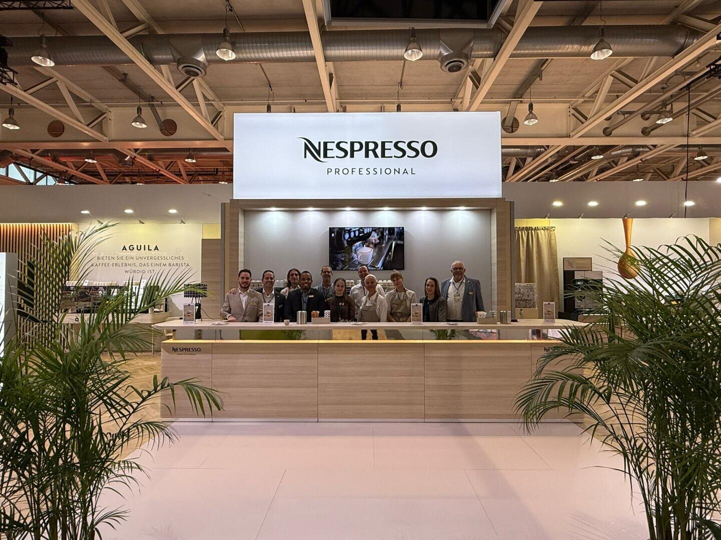 Eine Gruppe von Personen steht an einem Messestand hinter einer modernen Nespresso Professional-Theke, über ihnen ein großes Nespresso-Schild und im Vordergrund Grünpflanzen.