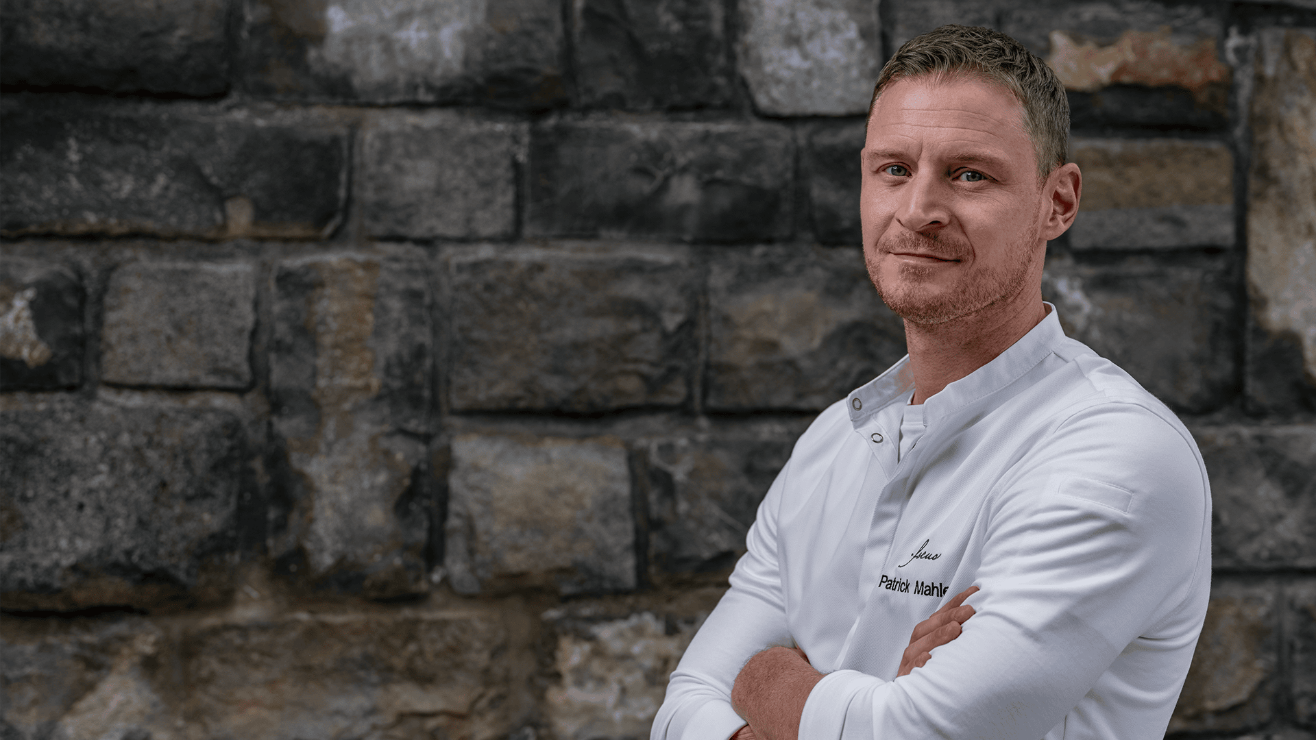 Un homme vêtu d'une veste de cuisine blanche se tient debout, les bras croisés et un léger sourire aux lèvres, devant un mur de pierre rustique.