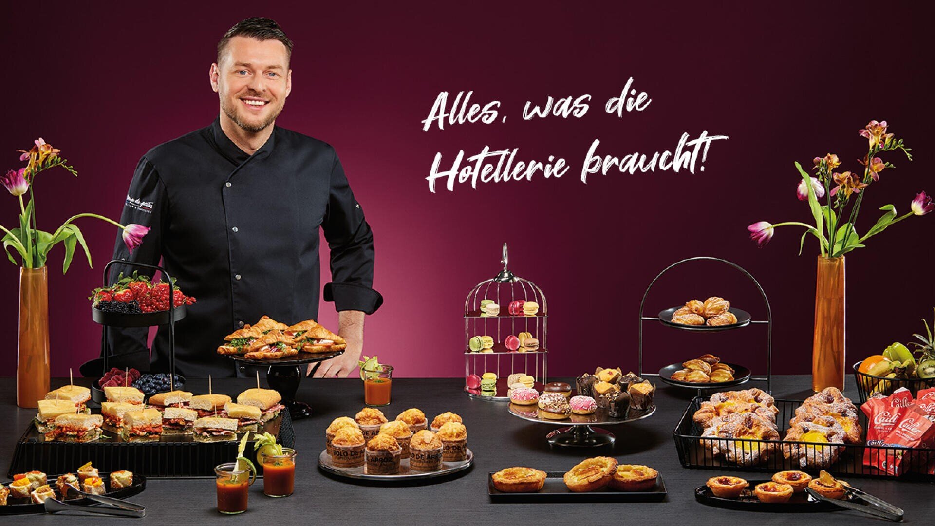 Ein lächelnder Koch in schwarzer Uniform steht hinter einem Tisch mit verschiedenen Backwaren, Sandwiches und Desserts. Bunte Blumen und Snacks schmücken den Tisch. Der deutsche Text an der Wand lautet: "Alles, was die Hotellerie braucht".