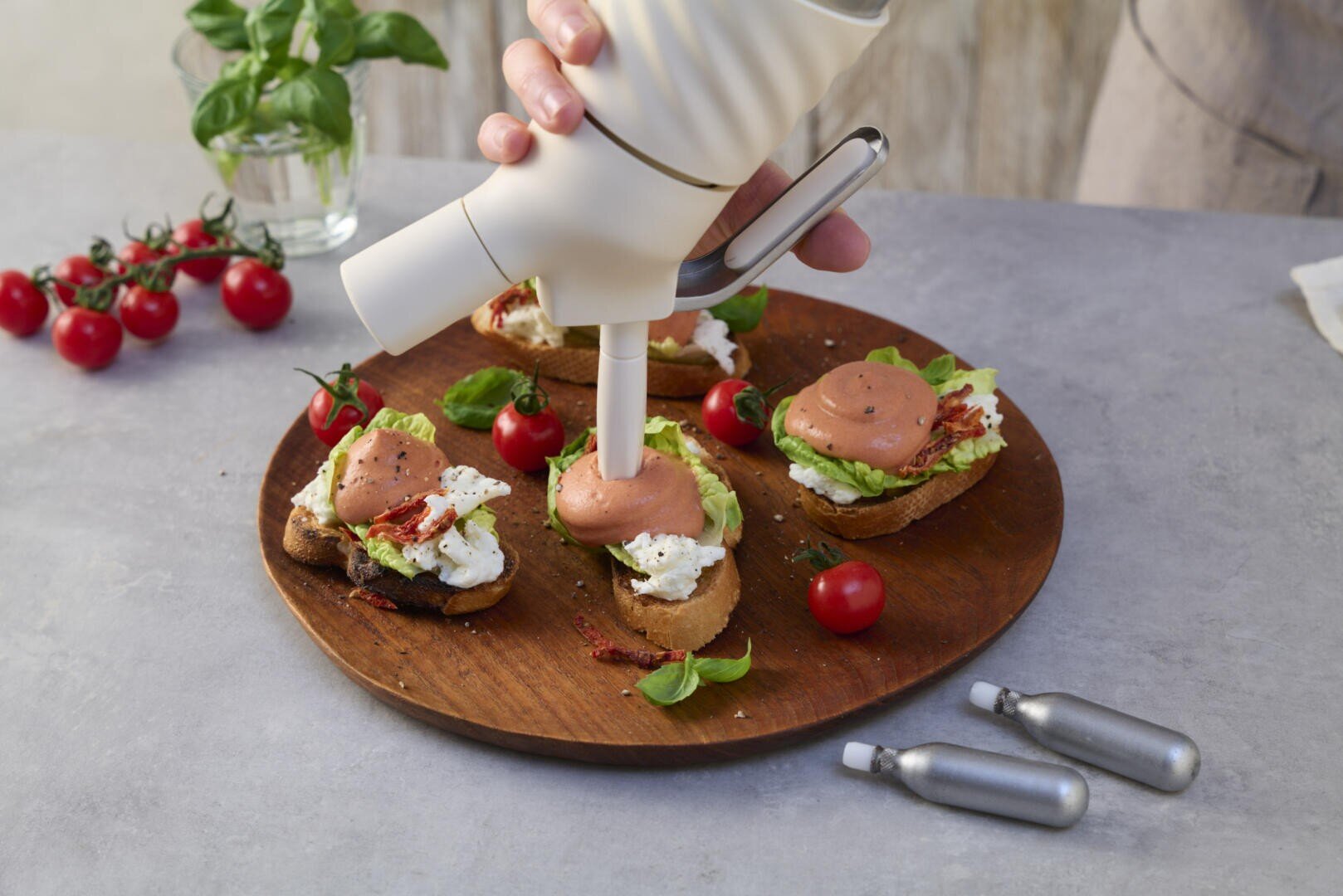 Eine Hand verwendet einen weißen Spender, um eine rosa Mousse auf geröstete Brotscheiben mit Käse, Avocado und Gemüse auf einem runden Holzbrett aufzutragen. Daneben stehen Kirschtomaten, Basilikum und Schlagsahne.