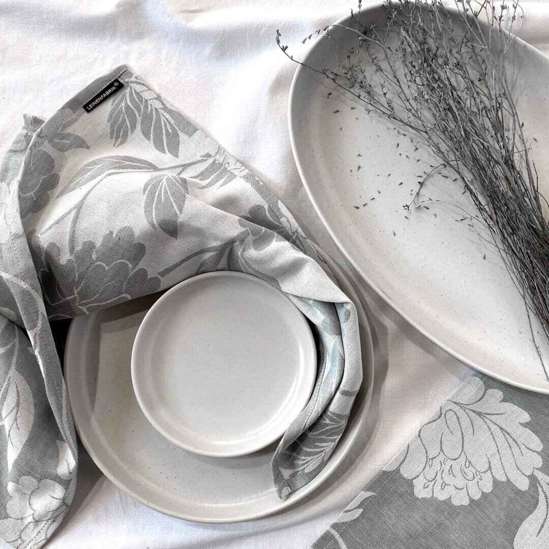 Ein minimalistisch gedeckter Tisch mit weißen Keramiktellern, einer kleinen Schale, einer grauen Serviette mit Blumenmuster und einer großen ovalen Platte mit getrockneten Zweigen, alles auf einem weißen Tischtuch angeordnet.