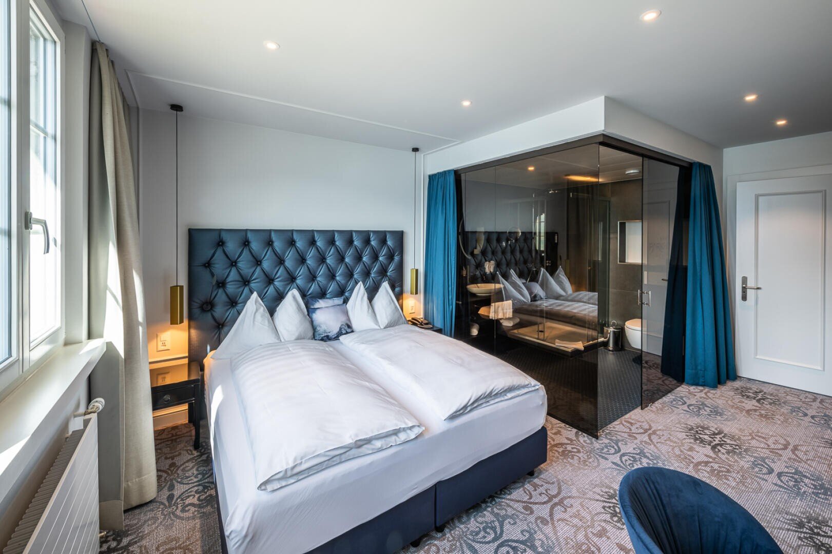 Ein modernes Hotelzimmer mit einem großen Bett, blauem Kopfteil, Nachttischen mit Lampen, blauen Vorhängen, einem gemusterten Teppich und einem Badezimmer mit Glaswänden. Natürliches Licht fällt durch ein Fenster auf der linken Seite ein.