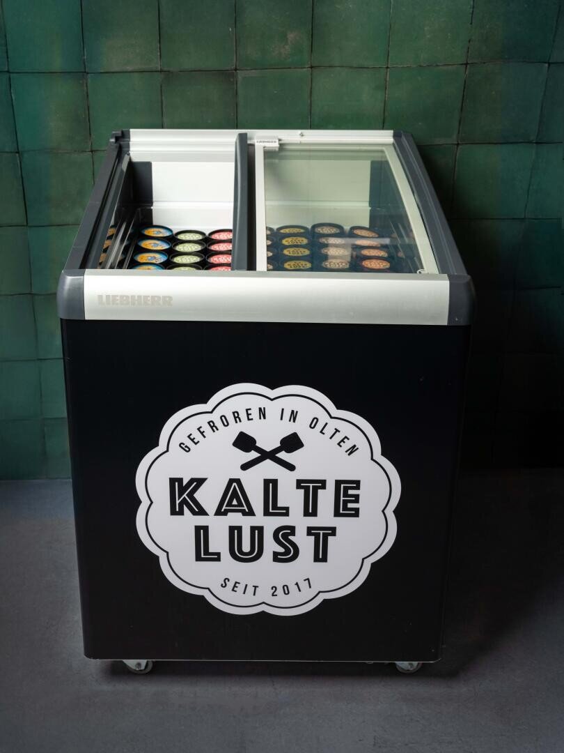 Ein schwarzer Eiscreme-Gefrierschrank mit Glasschiebetüren zeigt verschiedene Eiscremebecher. Die Vorderseite zeigt ein großes Kalte-Lust-Logo und einen deutschen Text. Die Gefriertruhe steht vor einer grün gekachelten Wand.