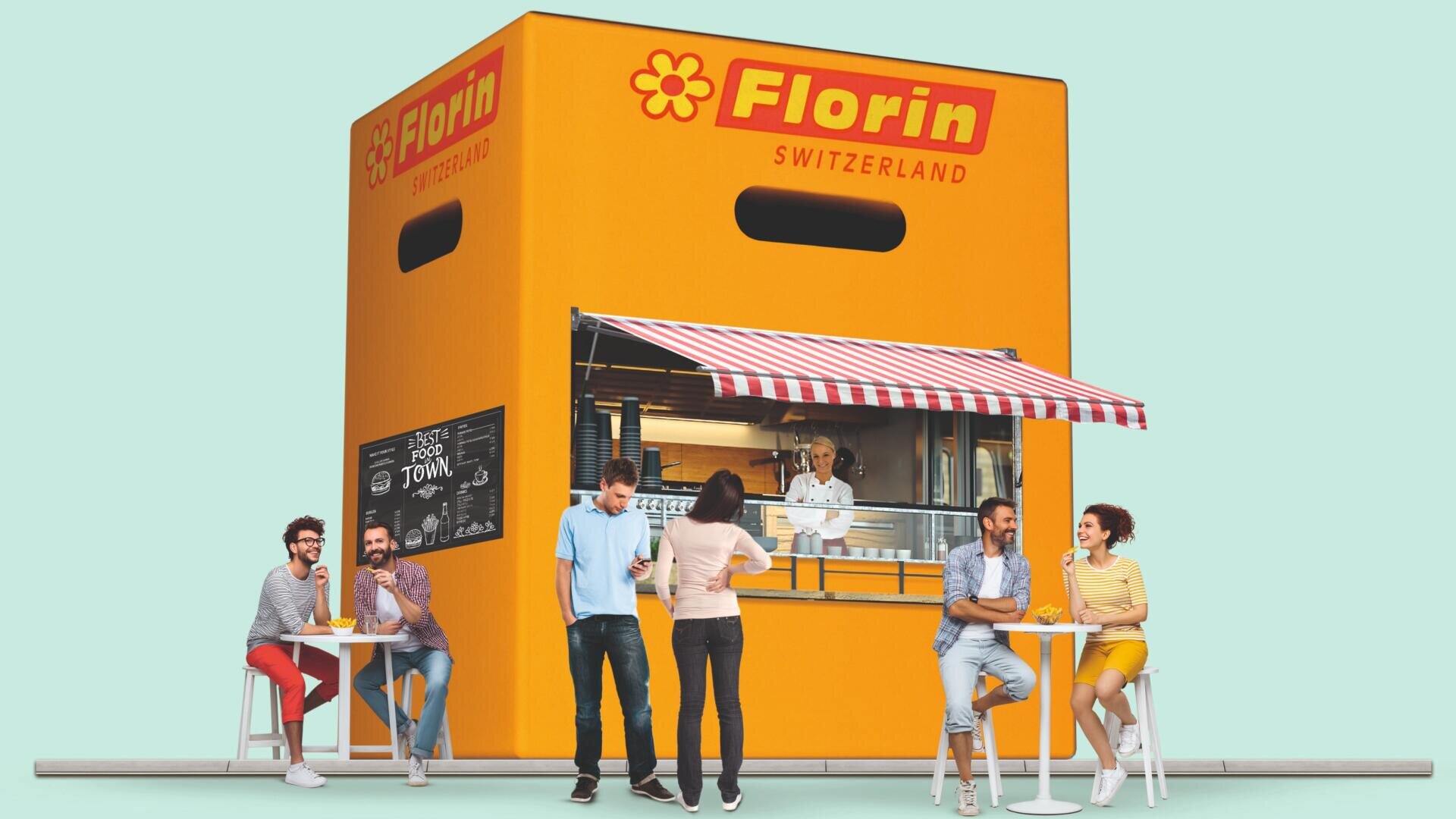 Ein gelber Lebensmittelkiosk in Form einer riesigen Florin-Schweiz-Box mit einer rot-weiß gestreiften Markise. An den weißen Tischen draußen wird gegessen, getrunken und geplaudert, während ein Angestellter vom Fenster aus Essen serviert.