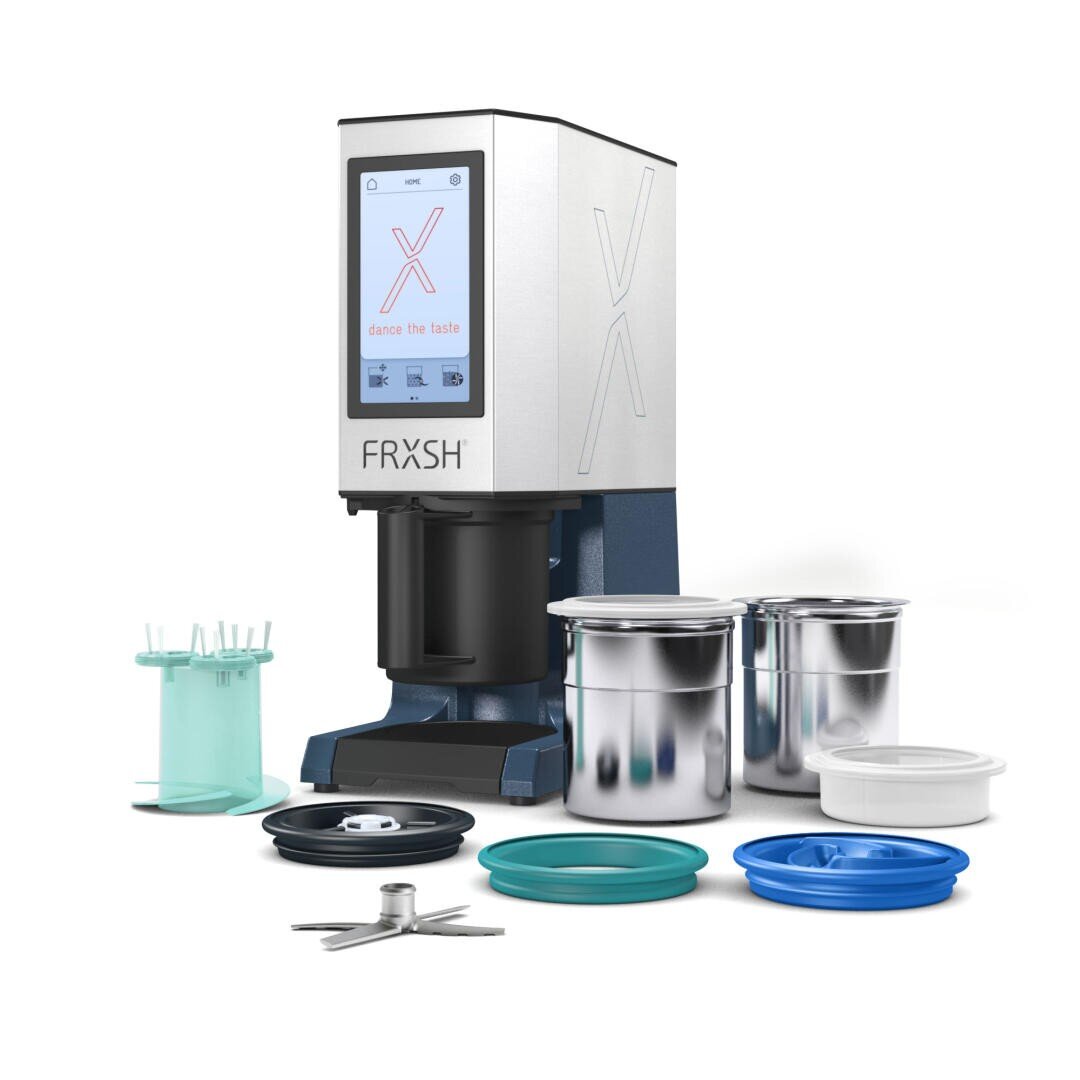 Un appareil de comptoir étiqueté FRXSH avec un écran tactile, deux récipients métalliques avec couvercles, des disques en plastique, une lame et d'autres accessoires disposés autour de l'appareil.