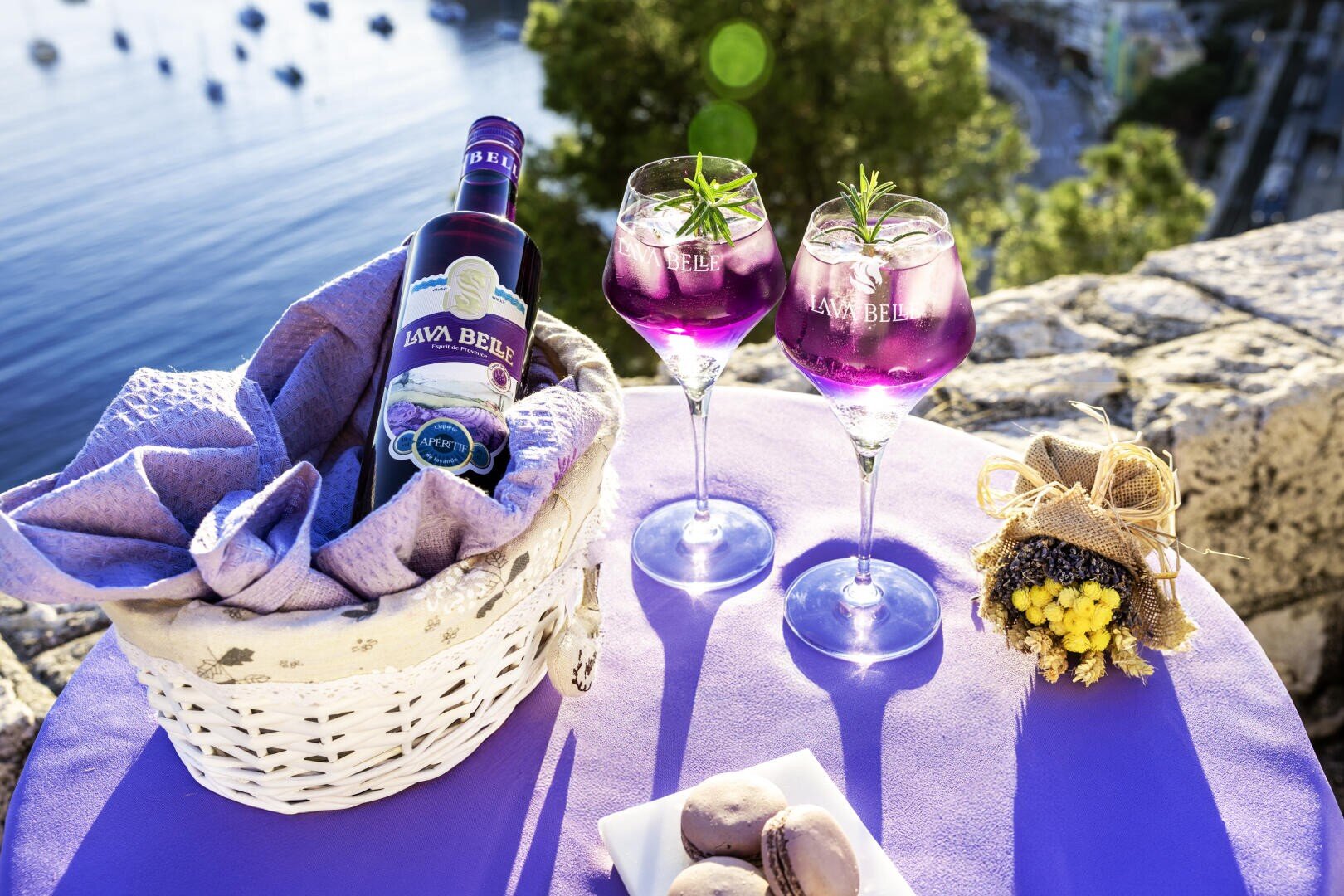 Une bouteille de la boisson violette Lavi Belle est posée dans un panier avec une serviette, à côté de deux verres remplis de la boisson et garnis de romarin, un petit bouquet de fleurs et une assiette de macarons sur une table violette avec vue sur l'eau et les bateaux.