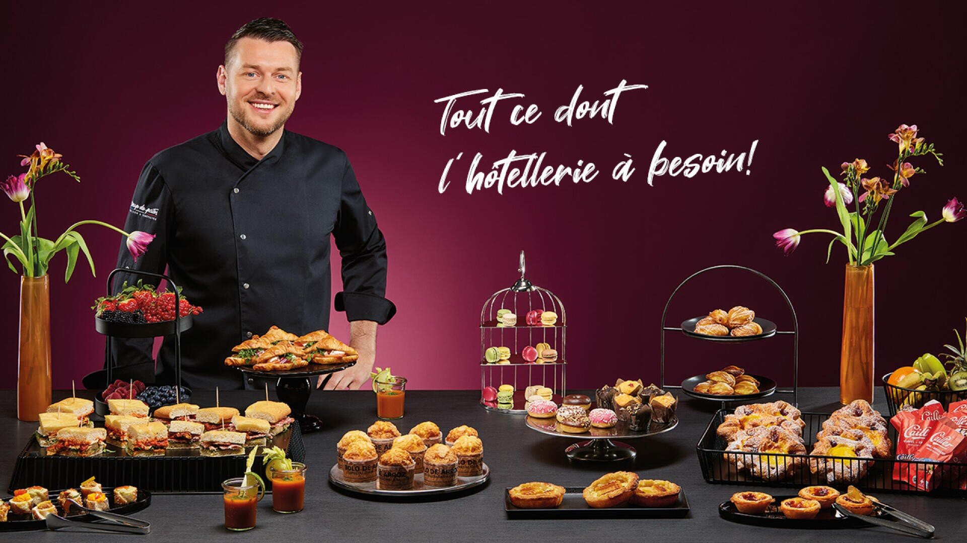 Un chef souriant en uniforme noir se tient derrière une table remplie de pâtisseries et de desserts assortis. Des fleurs décoratives et d'élégants plateaux de service sont disposés sur la table. Une phrase en français est écrite sur le fond marron derrière lui.