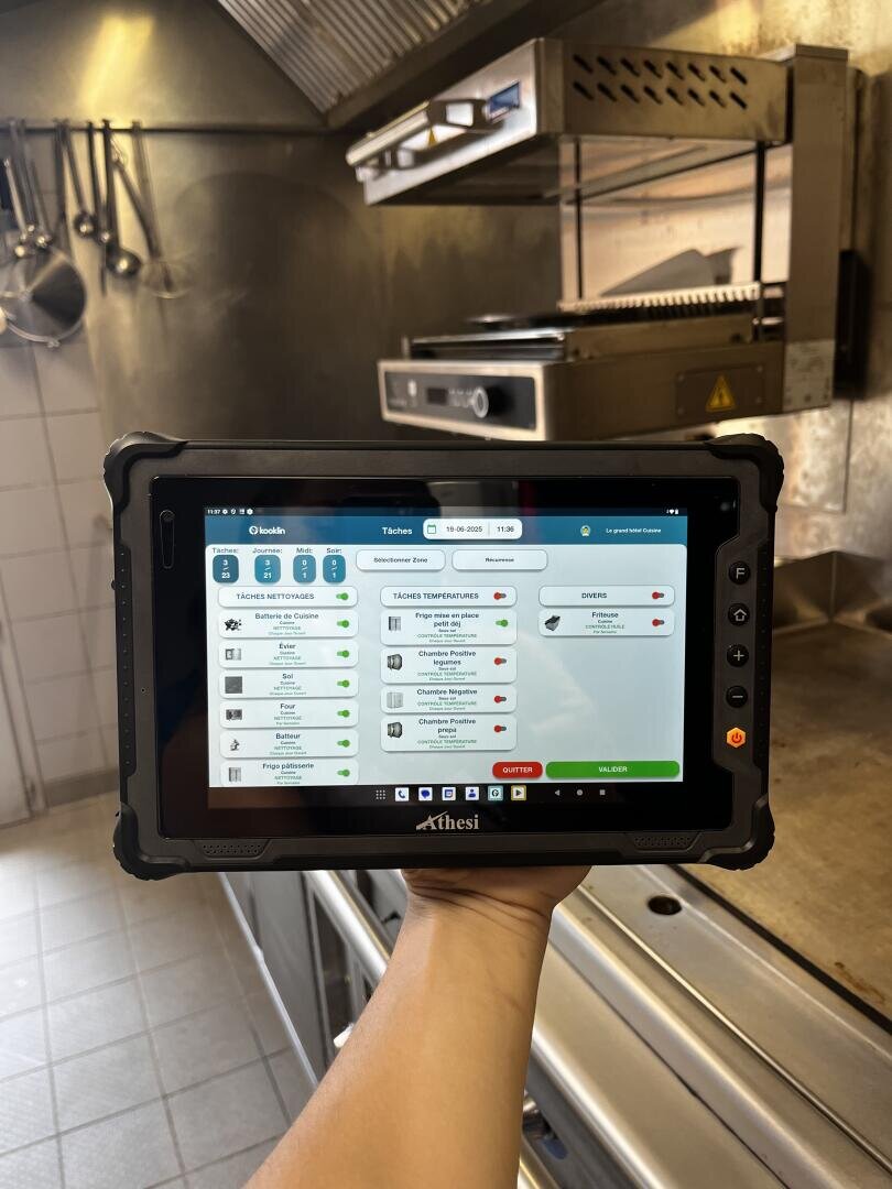 Une personne tient une tablette robuste sur laquelle s'affiche un logiciel de gestion de cuisine, dans une cuisine professionnelle en acier inoxydable avec des ustensiles suspendus et des appareils de cuisson en arrière-plan.