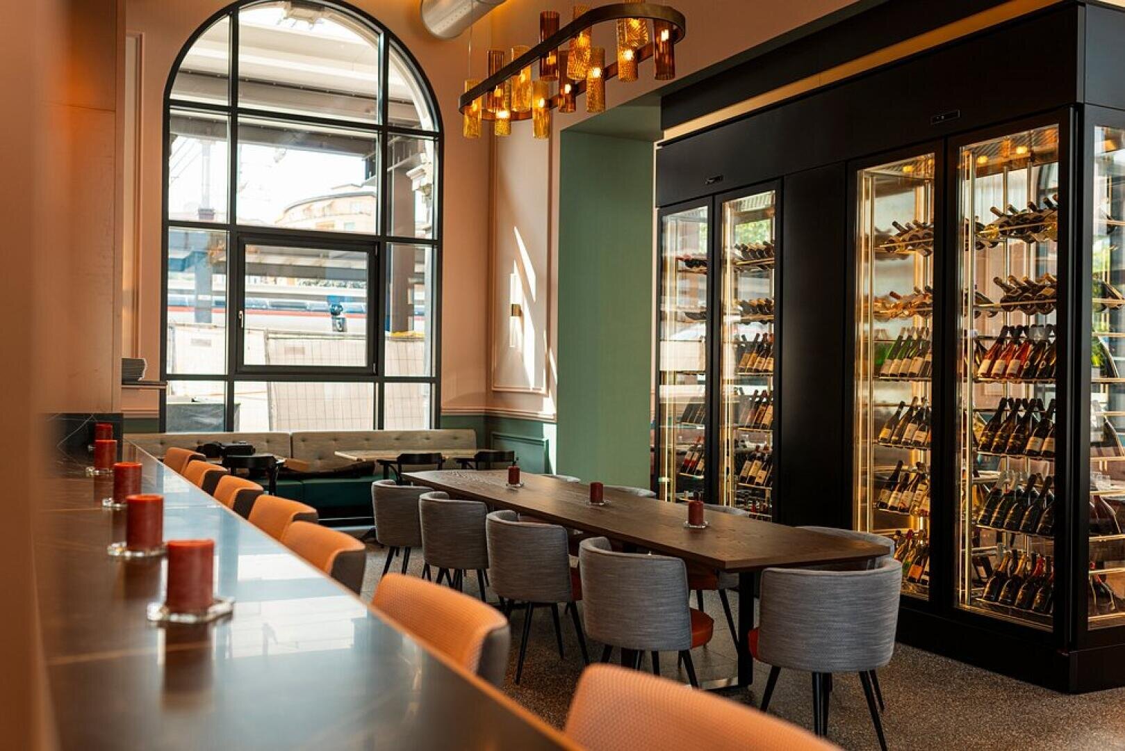 Un intérieur de restaurant élégant avec une longue table en bois entourée de chaises rembourrées, un mur de bouteilles de vin dans des armoires en verre, une rangée de bougies sur un comptoir et de grandes fenêtres cintrées laissant entrer la lumière naturelle.