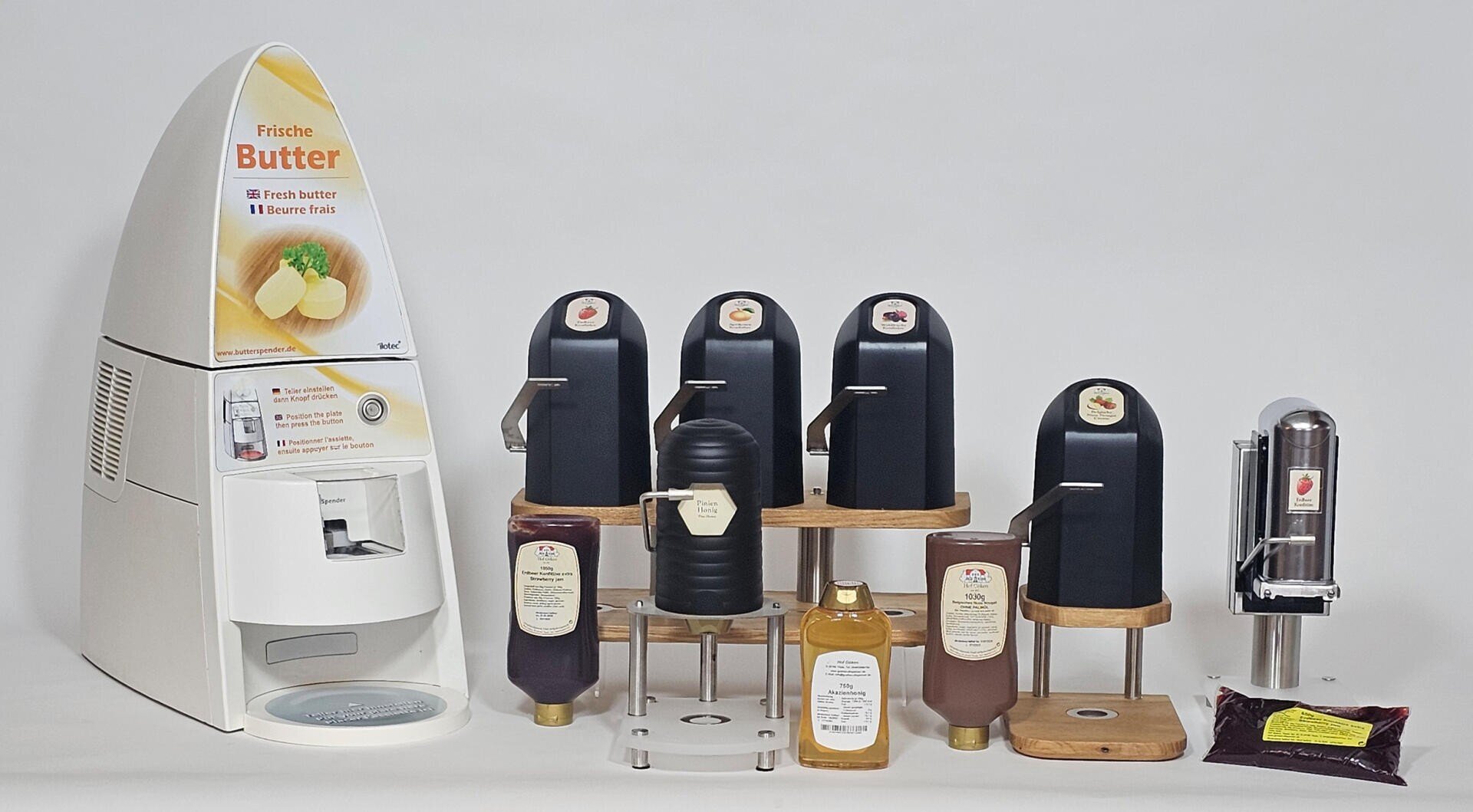 Eine Arbeitsplatte mit einem Butterspender auf der linken Seite und mehreren Gewürzspendern auf Ständern, die jeweils mit verschiedenen Soßen beschriftet sind. Außerdem sind Flaschen und eine Packung Sauce auf einem einfarbigen Hintergrund zu sehen.