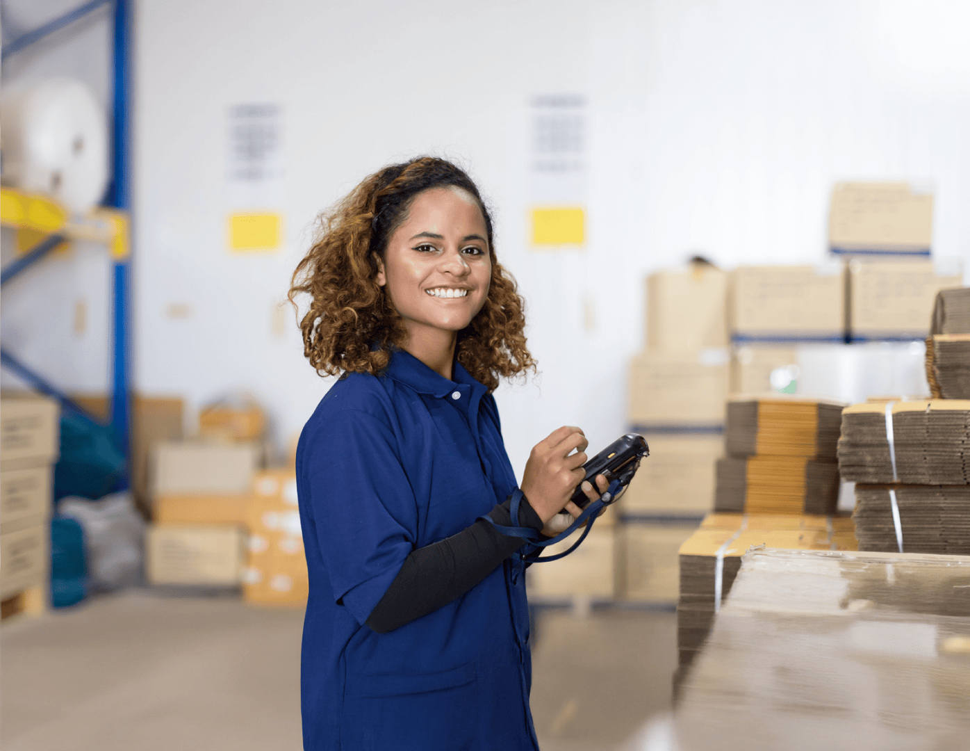Une femme vêtue d'un uniforme bleu se tient dans un entrepôt et sourit à la caméra en tenant un scanner à main. En arrière-plan, on voit des piles de cartons et de paquets.
