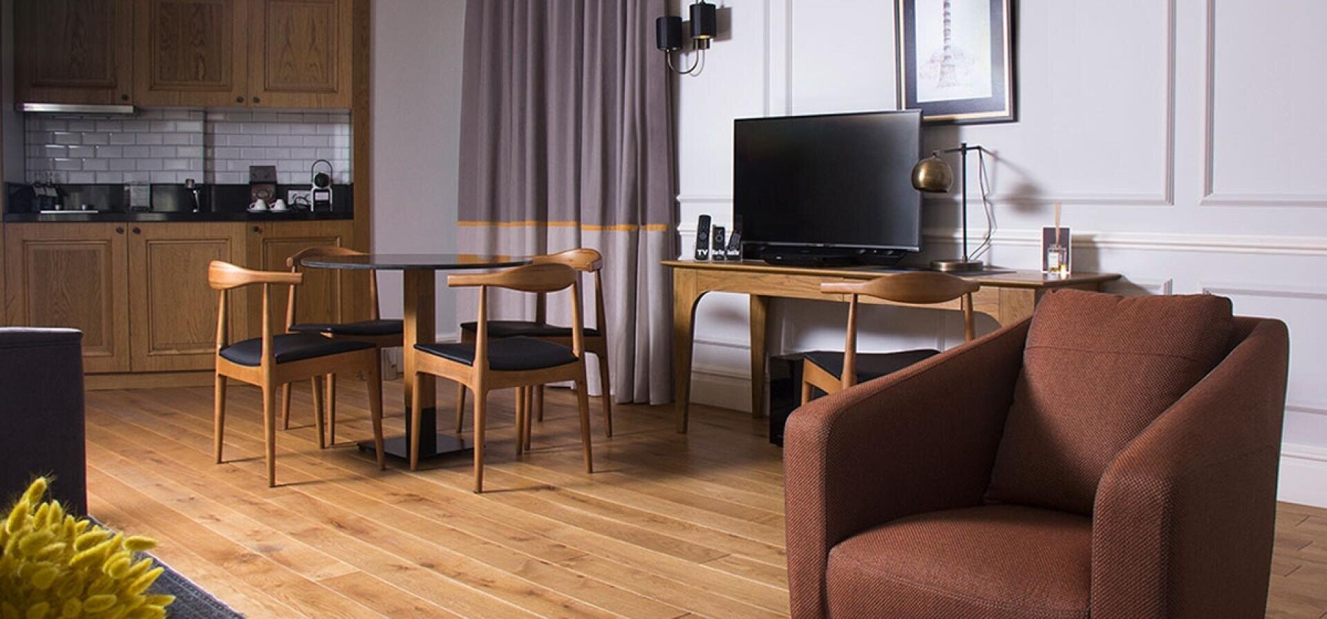 Moderner Wohnraum mit Holzfußboden, einem braunen Sessel, einem runden Esstisch mit vier Stühlen, einem Flachbildfernseher auf einem Holztisch und einer Küchenzeile im Hintergrund. Neutrale Töne und minimalistisches Dekor.