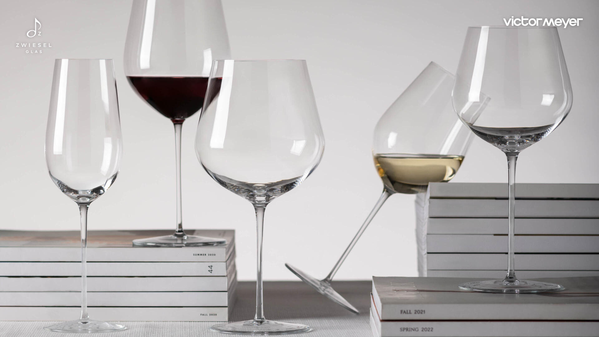 Vier Weingläser - zwei mit Rotwein, eines mit Weißwein und ein leeres - sind auf und neben gestapelten Büchern angeordnet. Der Hintergrund ist hellgrau, und in den Ecken sind die Logos Zwiesel Glas und victor meyer zu sehen.