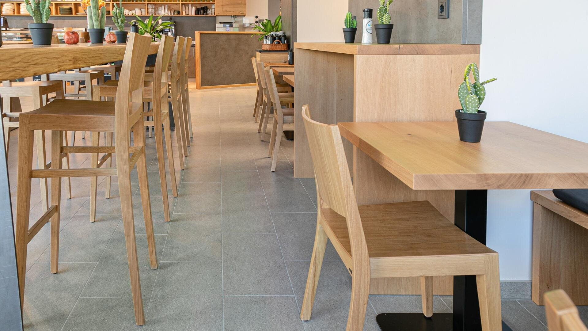 Modernes Café-Interieur mit hellen Holztischen und -stühlen, grauem Fliesenboden und kleinen Topfkakteen auf den Tischen. Der Raum ist hell und aufgeräumt und bietet eine gemütliche, minimalistische Atmosphäre.