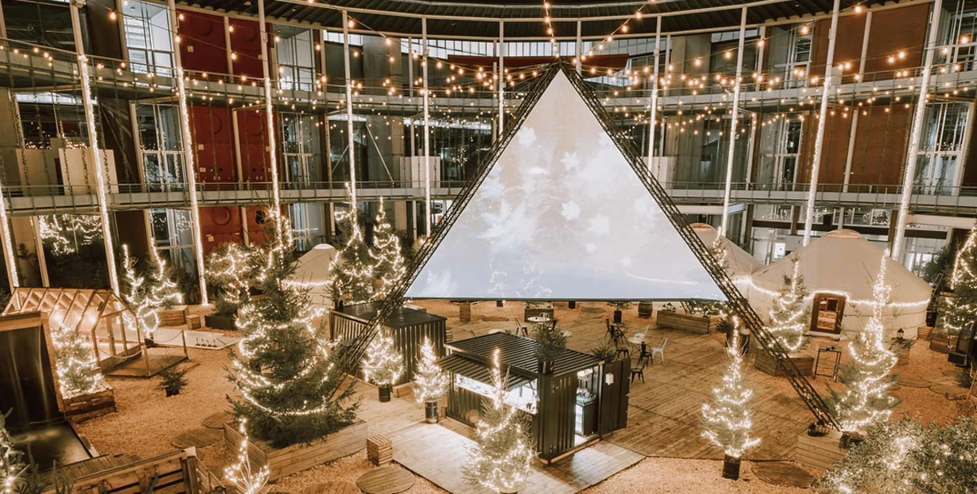 Ein festlich geschmückter Innenhof mit Lichterketten, beleuchteten Bäumen, einem großen dreieckigen Bildschirm in der Mitte und kleinen Strukturen und Sitzgelegenheiten, die über den Holzboden verstreut sind.