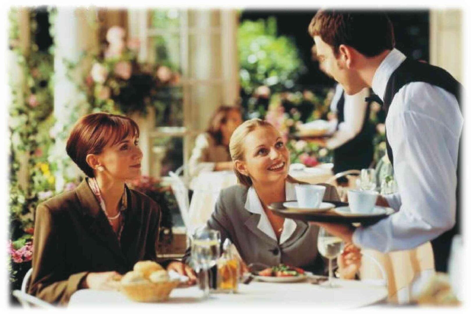 Deux femmes en tenue professionnelle sont assises à une table de restaurant, souriantes et regardant un serveur qui leur sert du café. Le décor est lumineux et élégant, avec des fleurs et d'autres convives en arrière-plan.