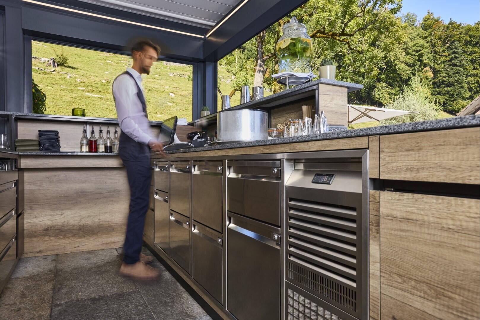 Ein Mann in formeller Kleidung arbeitet in einer modernen Außenküche mit Geräten aus rostfreiem Stahl, Holzschränken und einem großen Getränkespender vor einer Kulisse aus grünen Bäumen und grasbewachsenen Hügeln.