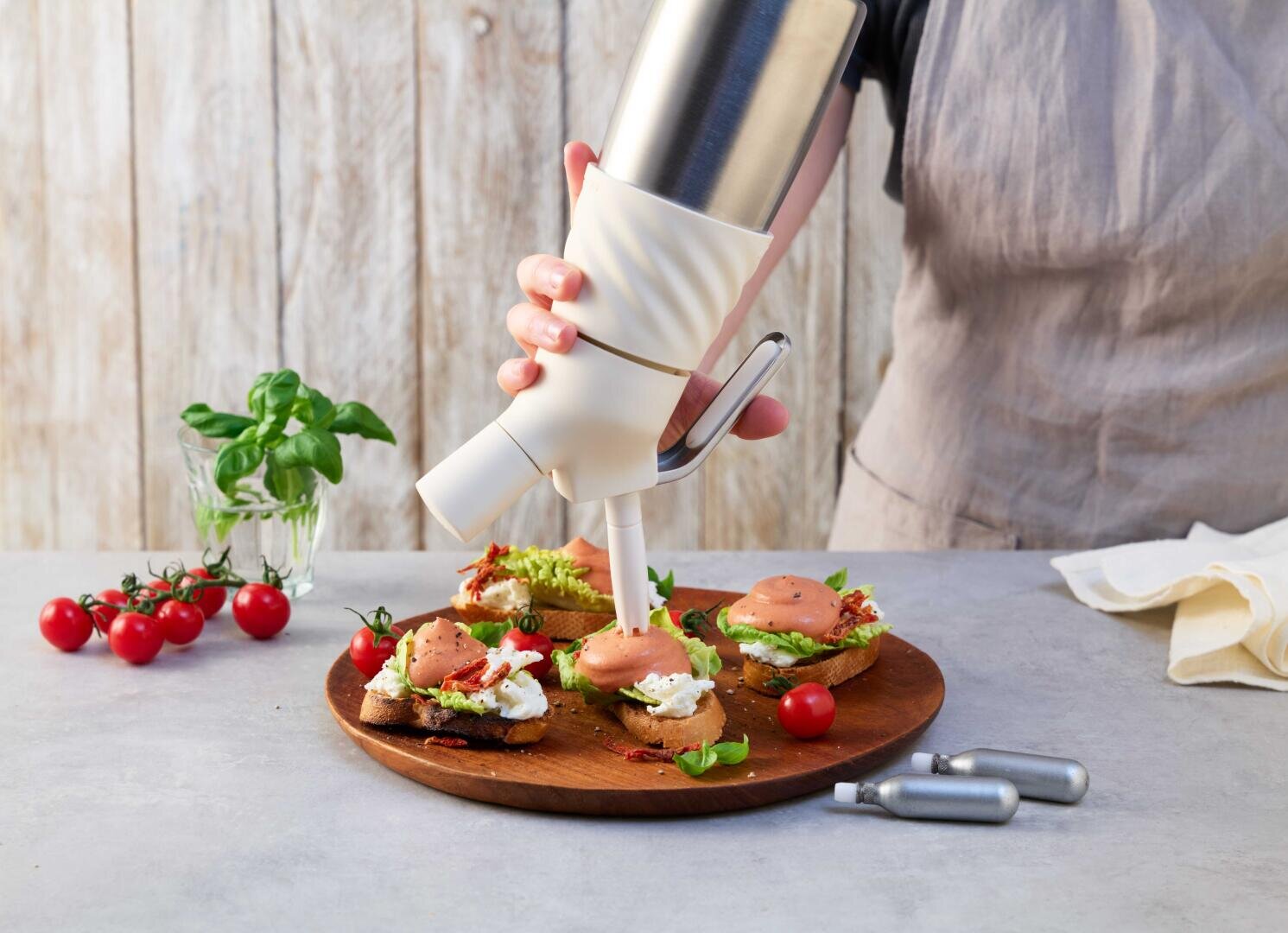 Eine Person verwendet einen Sahnespender, um Bruschetta auf einem Holzbrett zu belegen. Kirschtomaten, frisches Basilikum und Küchenutensilien liegen in der Nähe auf einer hellen Arbeitsplatte.