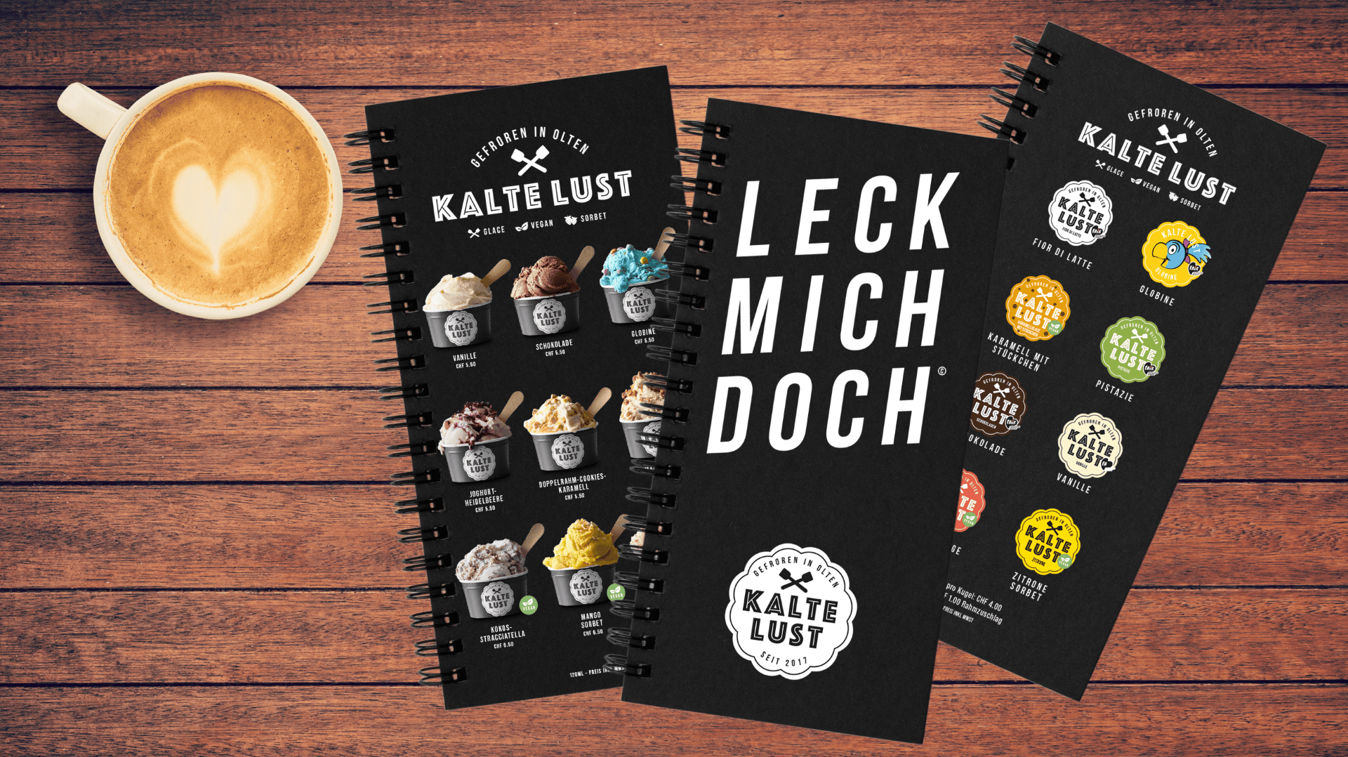 Eine Tasse Kaffee mit Latte Art steht neben drei spiralgebundenen Eiskarten der Marke Kalte Lust, die auf einem Holztisch ausgestellt sind. Die Karten zeigen Bilder und Geschmacksrichtungen von Eiscreme sowie den fettgedruckten Text „LECK MICH DOCH“.