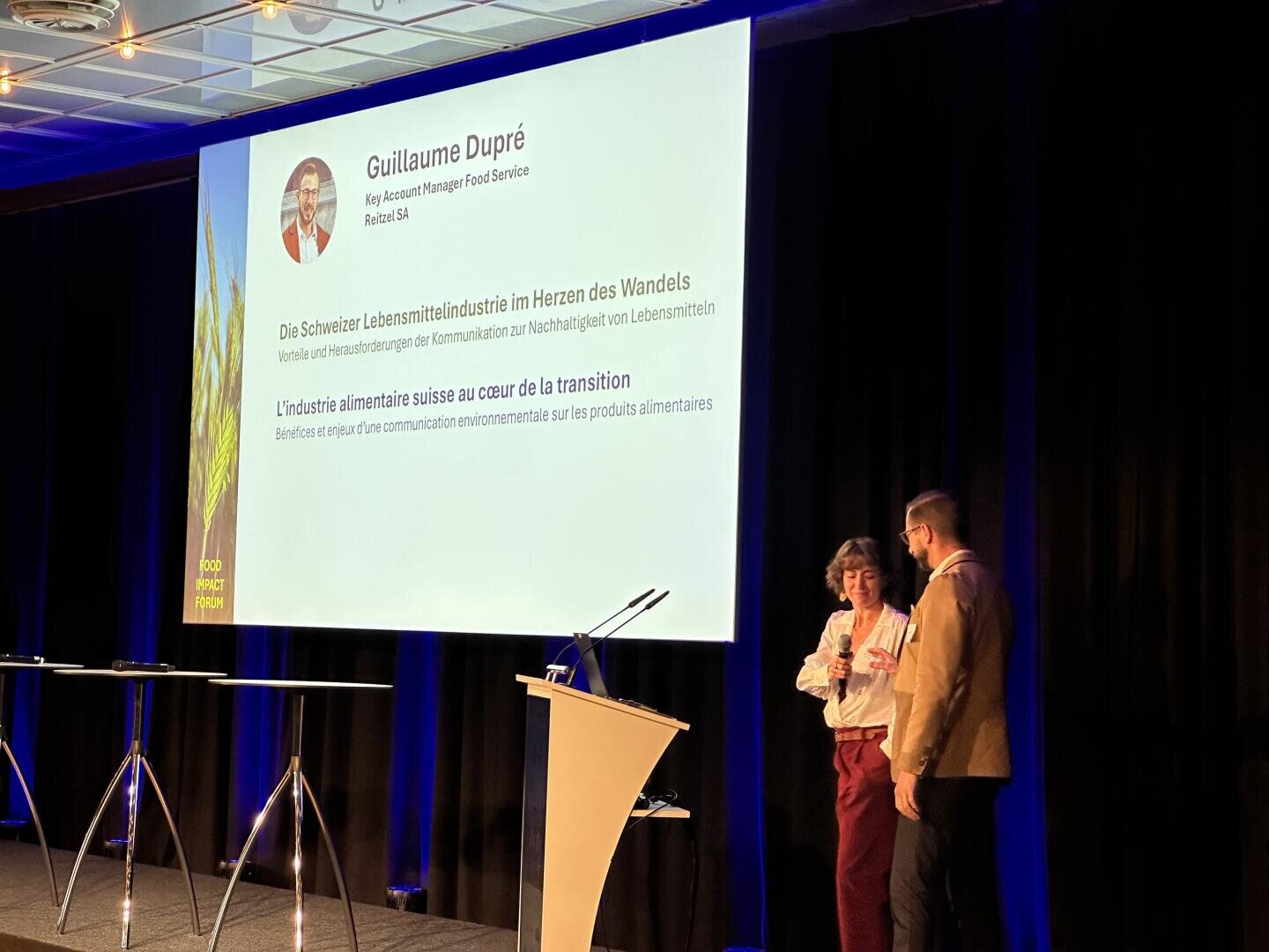 Zwei Personen stehen auf einer Bühne unter Scheinwerfern und sprechen in der Nähe eines Podiums. Auf einem großen Bildschirm hinter ihnen wird eine Präsentationsfolie mit einem Foto und Details zu Guillaume Dupré und seinem Vortrag über die Schweizer Lebensmittelindustrie gezeigt.