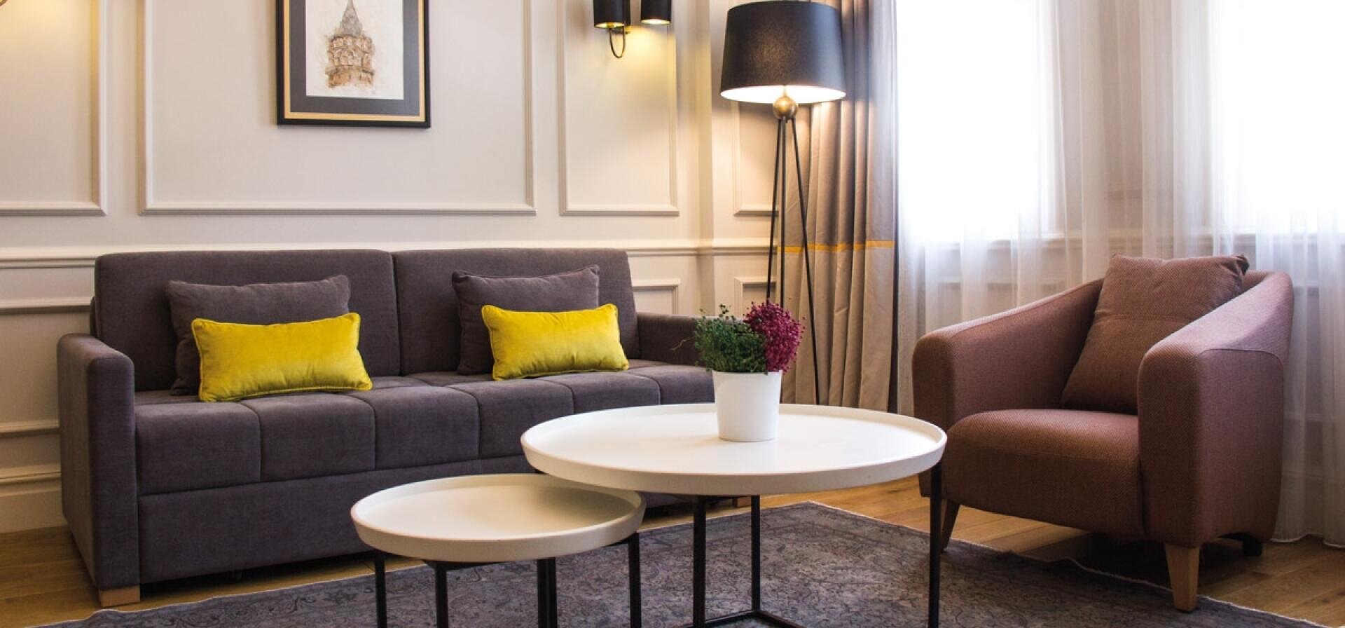 Ein modernes Wohnzimmer mit einem grauen Sofa, zwei gelben Kissen, einem braunen Sessel, zwei runden weißen Couchtischen mit einer Topfpflanze, einer Stehlampe, gerahmter Kunst, Wandleuchten und durchsichtigen Vorhängen vor einem großen Fenster.