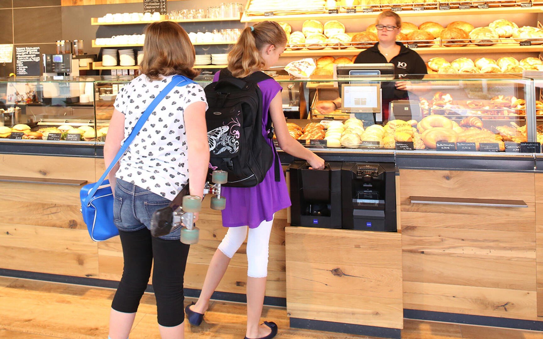 Zwei Mädchen stehen an der Theke einer Bäckerei; die eine wählt etwas an einem Selbstbedienungsautomaten aus, während die andere ein Skateboard hält. Hinter der Theke steht eine Mitarbeiterin vor Regalen mit Brot und Gebäck.