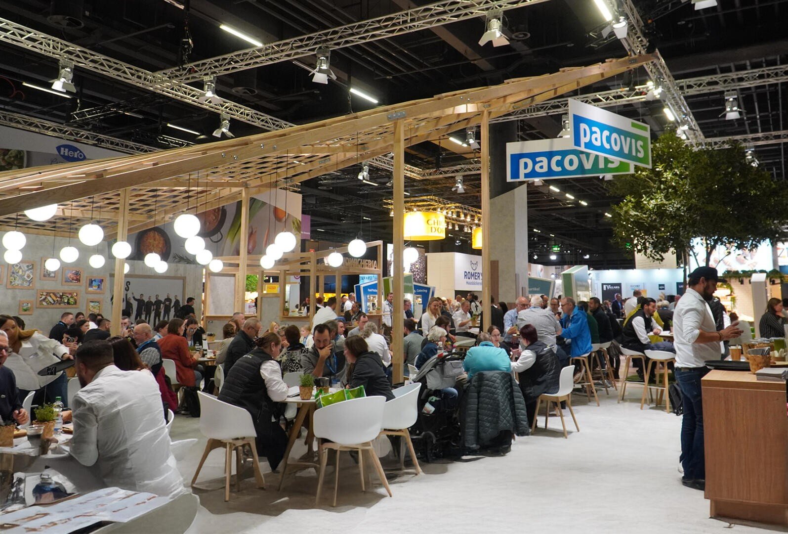 Une foire ou une exposition couverte et animée où de nombreuses personnes sont assises à des tables, mangent et discutent. Des lumières vives pendent du plafond et un panneau pacovis est visible au-dessus de la foule. Des stands et des étalages remplissent l'arrière-plan.