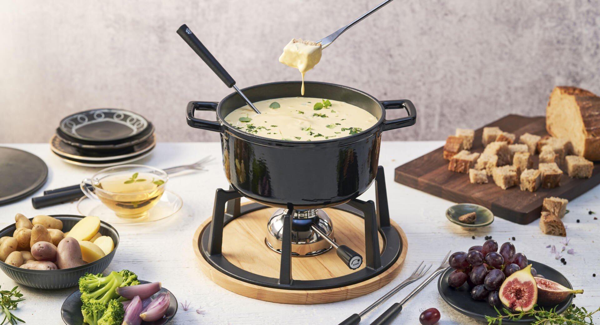 Ein schwarzer Fonduetopf mit geschmolzenem Käse steht auf einem Brenner, umgeben von Tellern, Brotwürfeln, Weintrauben, Kartoffeln, Brokkoli, Schalotten und Kräutern. Zwei Gabeln halten in Käse getauchtes Brot über dem Topf.