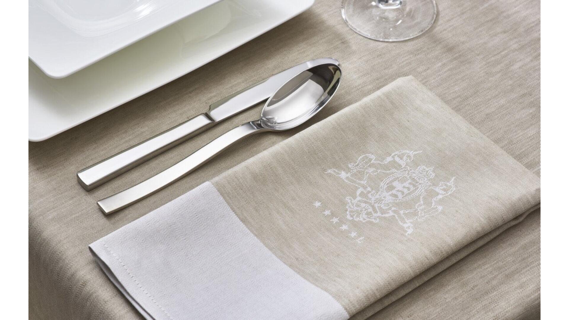 Gros plan sur une table à manger soigneusement dressée, avec une fourchette et une cuillère sur une serviette beige brodée d'un écusson blanc, à côté d'une assiette blanche et d'un verre à vin sur une nappe beige.