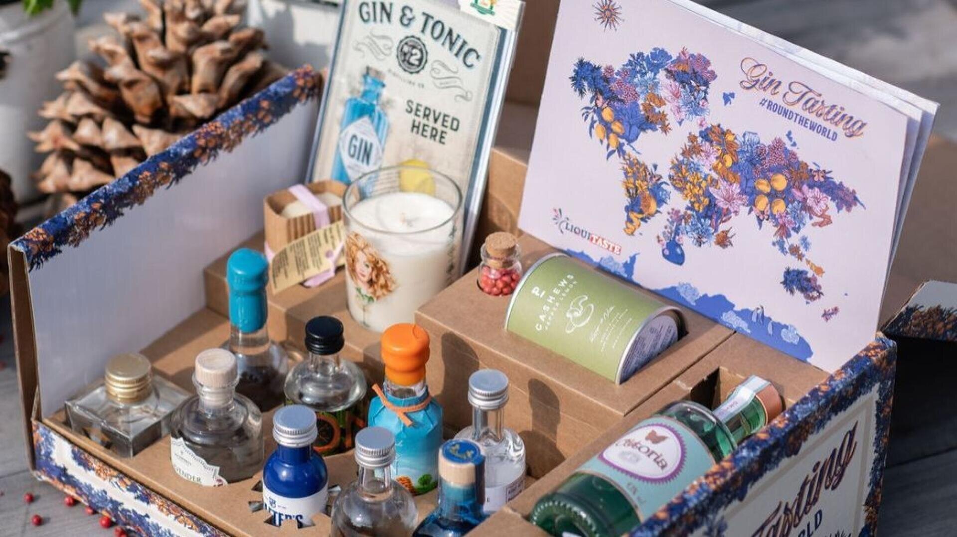 Ein offener Geschenkkarton mit Mini-Gin-Flaschen, Tonic-Dosen, einer Kerze, einem Büchlein mit der Aufschrift "Gin Tasting", einem Tannenzapfen und verschiedenen Dekorationsartikeln, die alle für eine Gin-Verkostung arrangiert wurden.