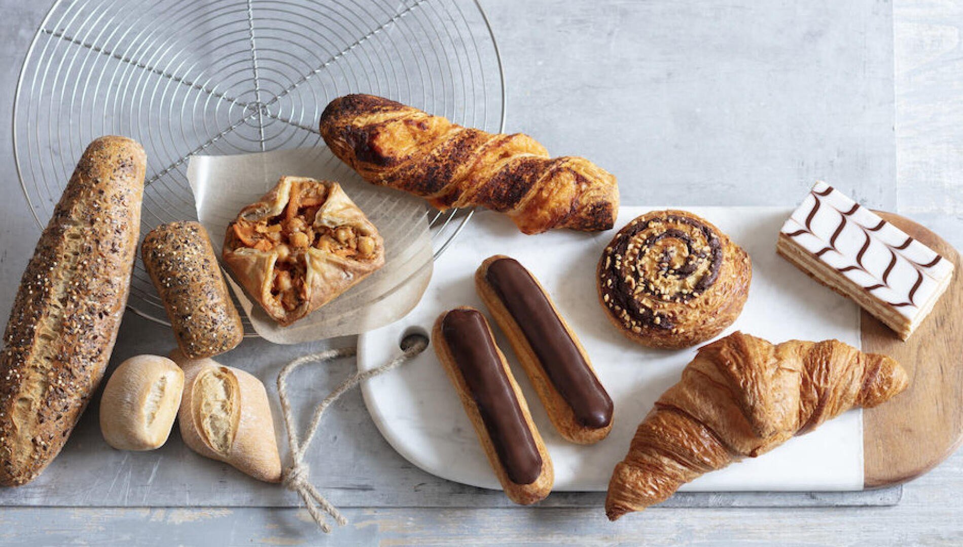 Eine Auswahl an Backwaren, darunter Baguette, Körnerbrot, Croissants, Eclairs, eine Zimtrolle, ein Apfelkuchen und ein Stück Schichtdessert, angeordnet auf einem Marmor- und Holzbrett und einem Drahtgestell.