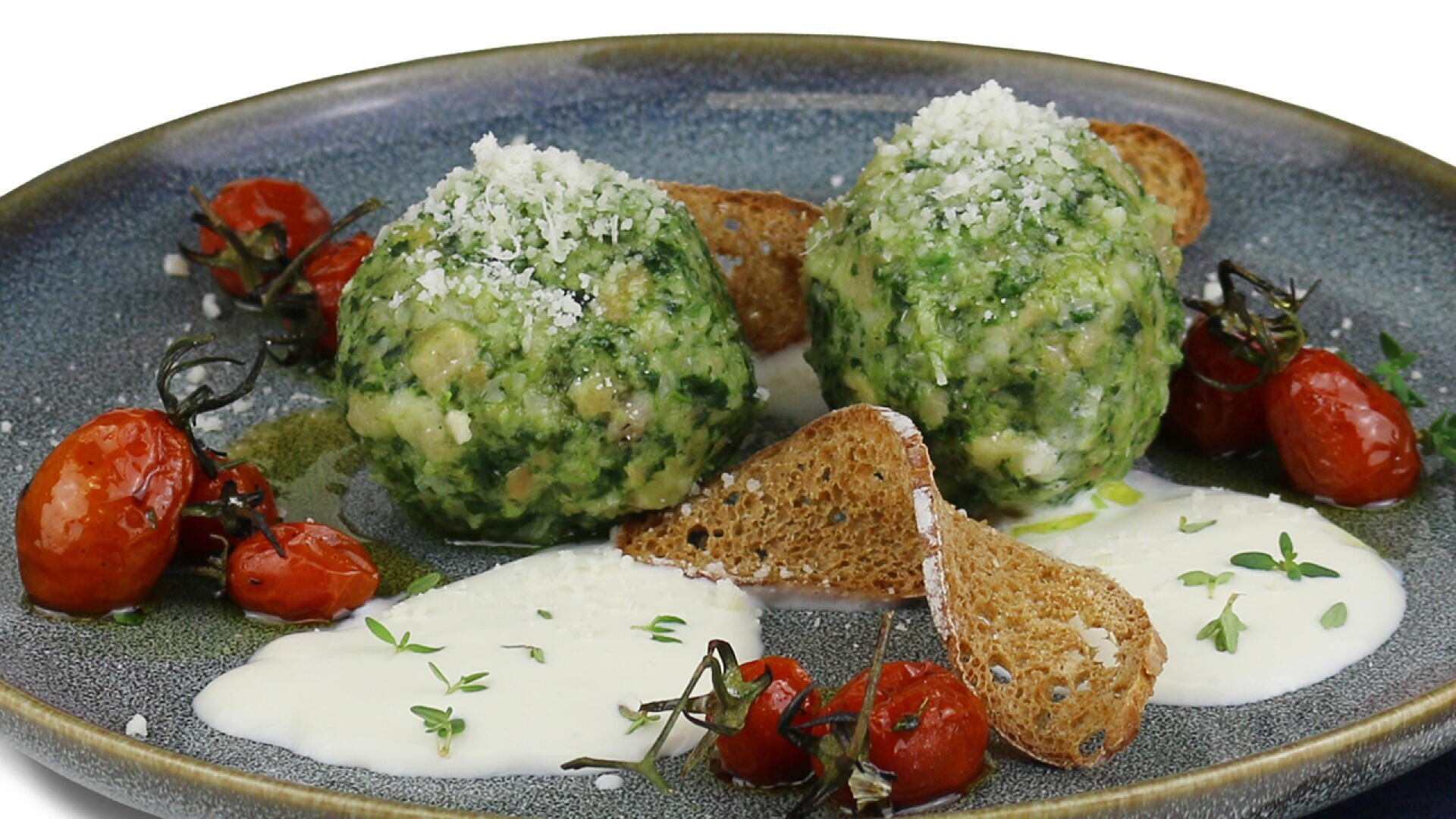 Zwei mit geriebenem Käse garnierte Spinat-Käse-Knödel liegen auf einem grauen Teller, umgeben von gerösteten Kirschtomaten, gerösteten Brotscheiben, weißer Soße und frischen Kräutern.