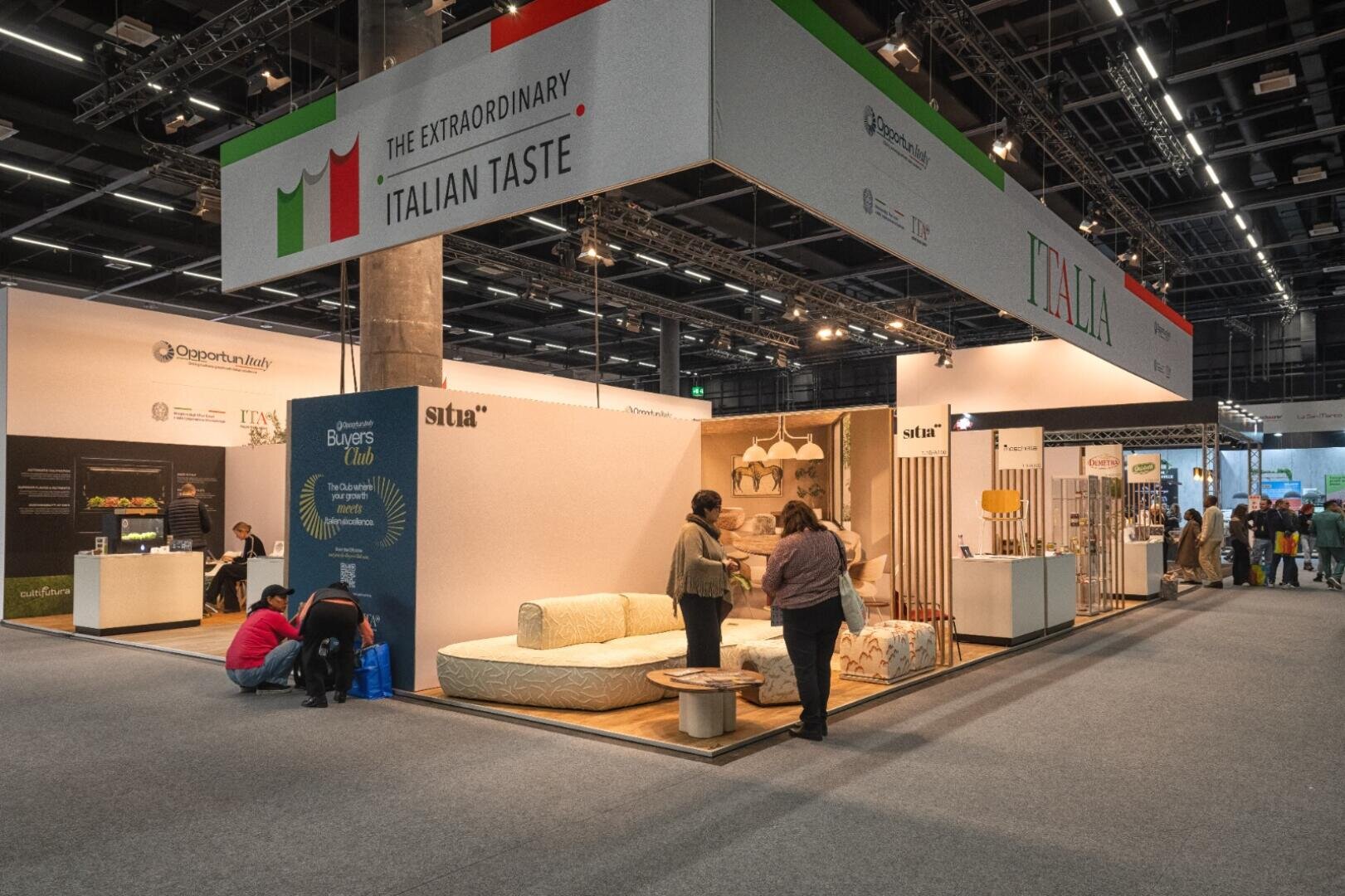 Un stand d'exposition avec un panneau portant l'inscription The Extraordinary Italian Taste. Lors d'un salon, des personnes sont debout ou assises près d'un étalage de meubles modernes, sous des lumières vives.