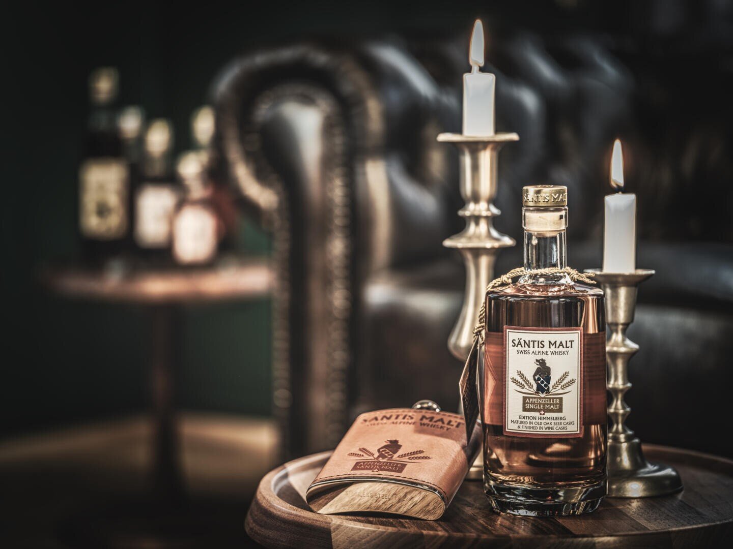 Eine Flasche Säntis Malt Whisky und ein Lederkolben stehen auf einem Holztisch neben brennenden Kerzen. Ein Ledersofa und weitere Flaschen verschwimmen im Hintergrund und schaffen eine gemütliche, elegante Atmosphäre.