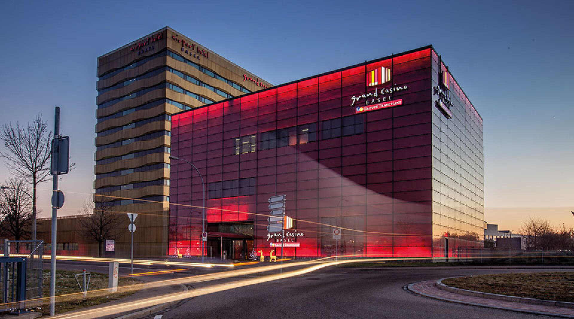 Das Grand Casino Basel mit seiner Glasfassade, die in der Abenddämmerung rot beleuchtet wird. Auf der Straße davor sind Lichtspuren von Autos zu sehen.