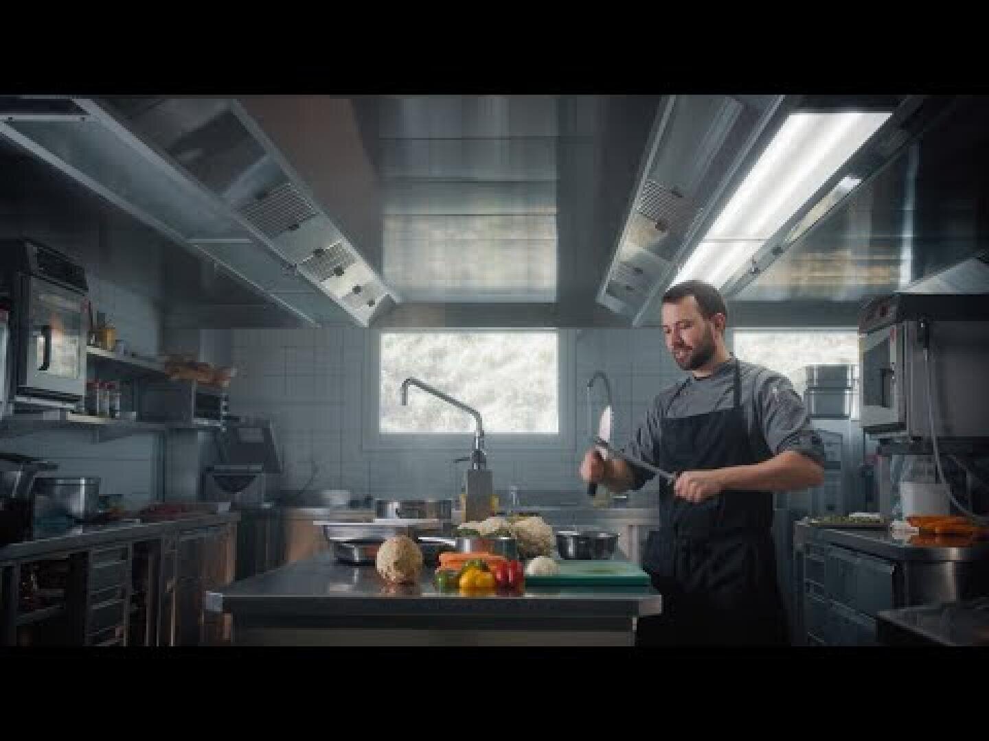 Ein Koch in einer modernen Küche schneidet Gemüse auf einem Tresen. Verschiedene Zutaten und Utensilien sind darauf verteilt, und durch ein Fenster im Hintergrund fällt Sonnenlicht ein.