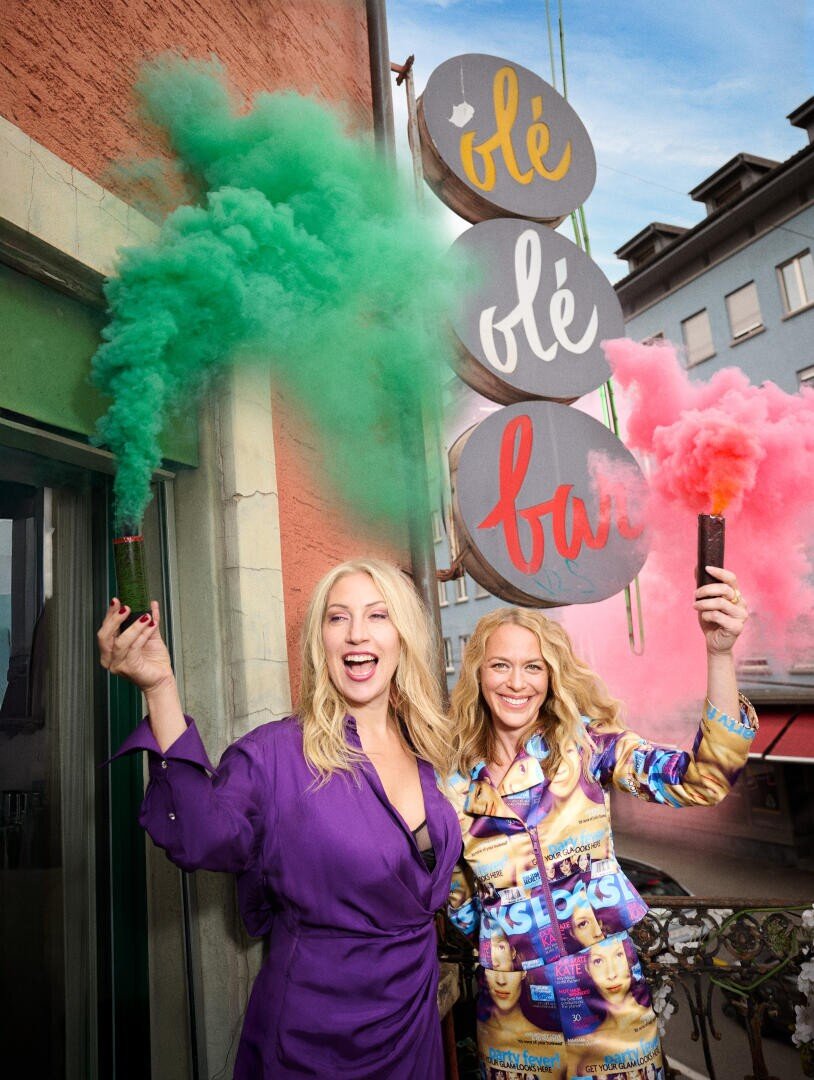 Zwei lächelnde Frauen stehen auf einem Balkon, jede hält eine Rauchfackel in der Hand - eine grün, eine rosa. Hinter ihnen steht ein rundes Schild mit der Aufschrift olé olé bar. Beide Frauen tragen bunte Kleidung, und im Hintergrund sind Gebäude zu sehen.