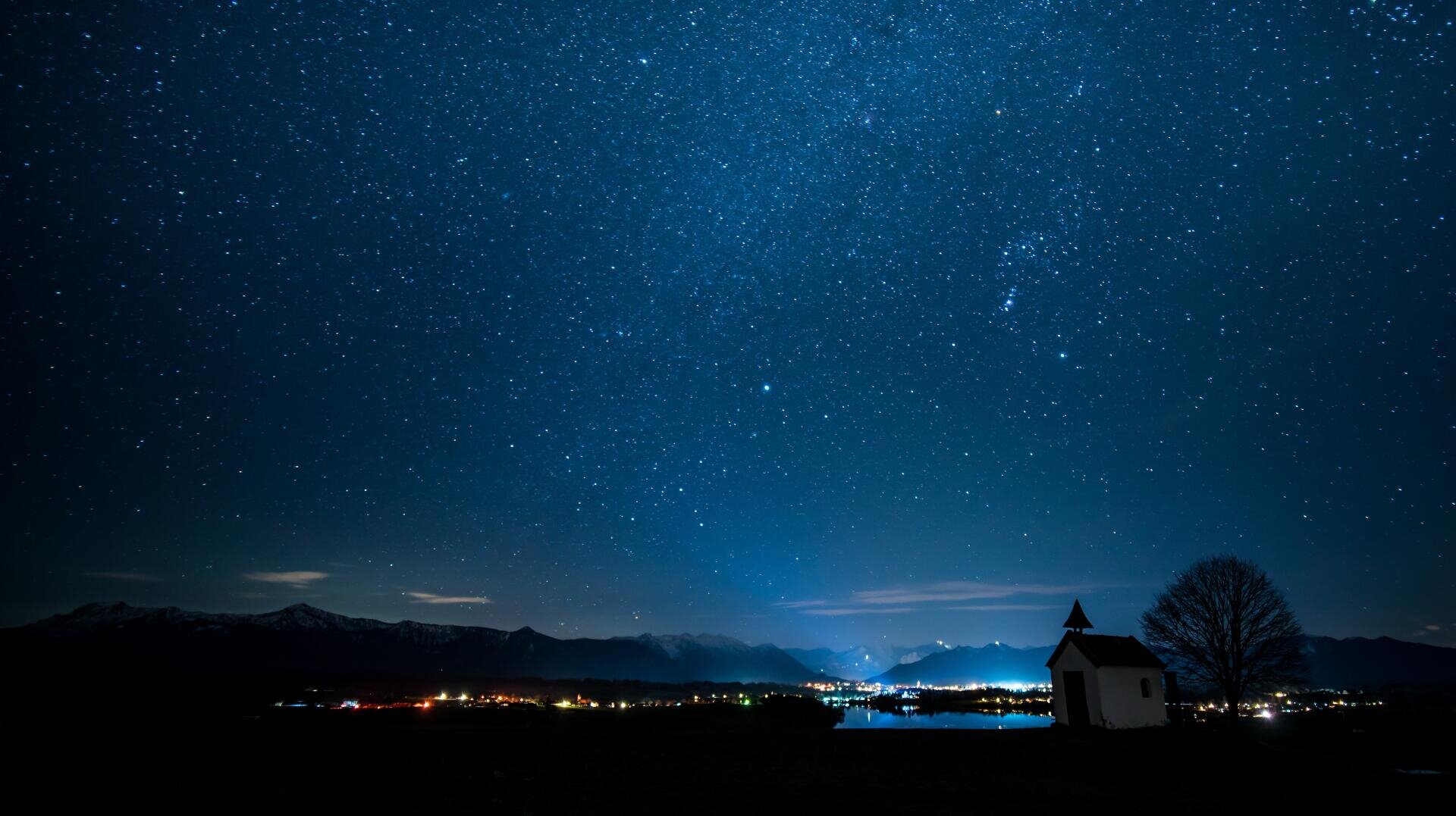 Eine kleine Kapelle und ein Baum heben sich von einem sternenübersäten Nachthimmel ab, während sich im Hintergrund die Lichter der Stadt, die Berge und ein ruhiges Gewässer abzeichnen.
