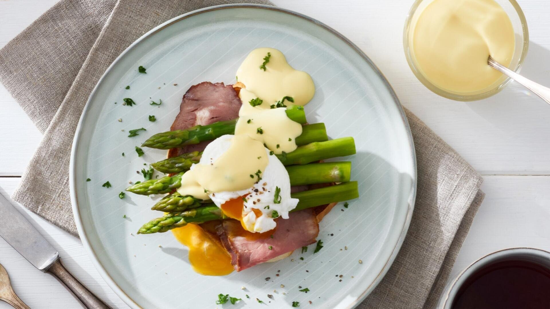 Ein Teller mit pochierten Eiern, Roastbeef, Spargel und Sauce Hollandaise, garniert mit gehackten Kräutern, steht auf einer hellen Serviette neben einer Gabel, einem Messer, einer Schüssel mit Sauce und einem Glas mit dunklem Getränk.