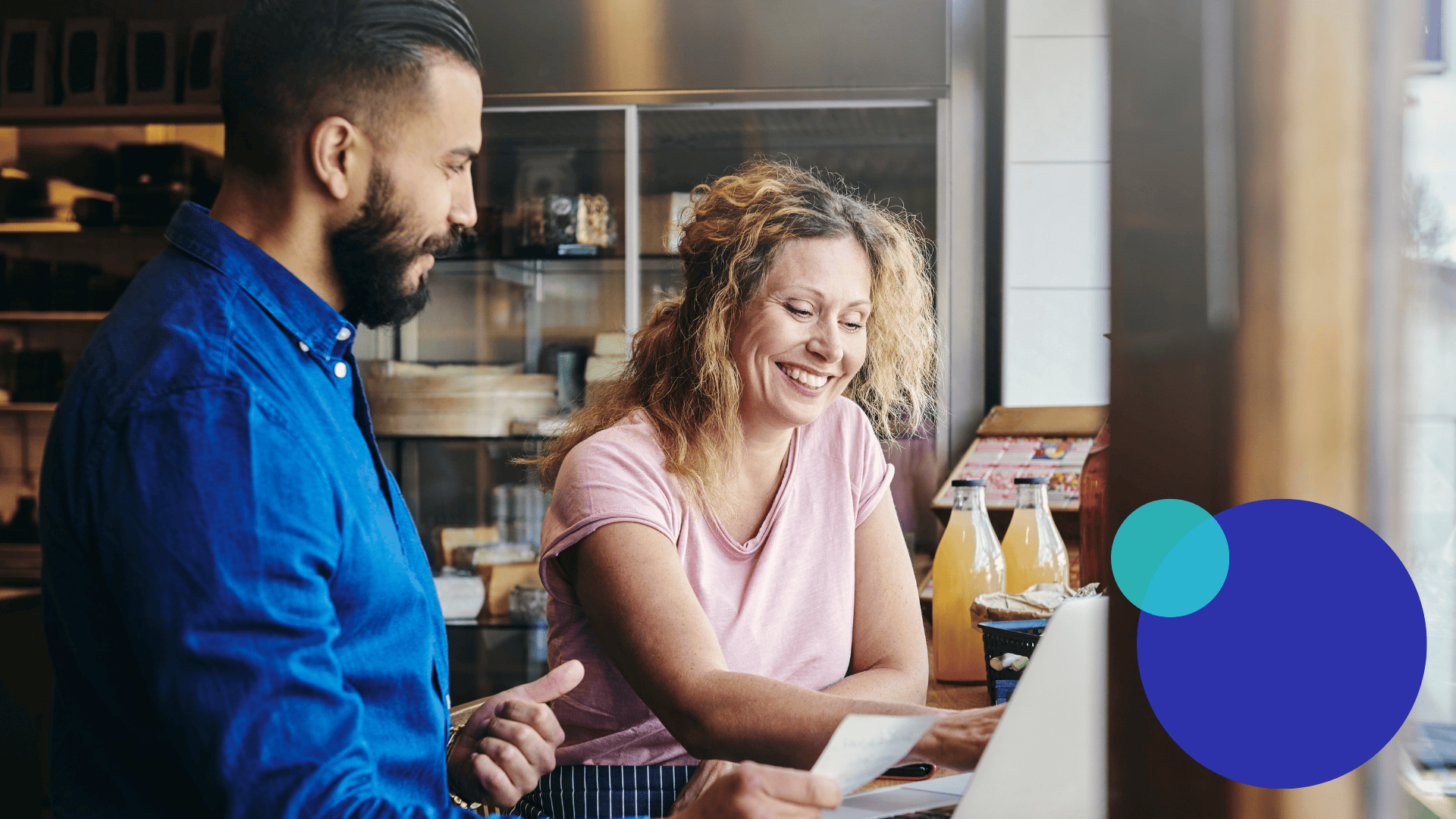 Ein Mann und eine Frau stehen an einem Tresen in einem hellen Café. Die Frau lächelt, während sie auf eine Quittung schaut, und der Mann gestikuliert, während sie sich unterhalten. Im Hintergrund sind Getränke in Flaschen und Gebäck zu sehen. Blaue grafische Formen überlagern die Ecke.