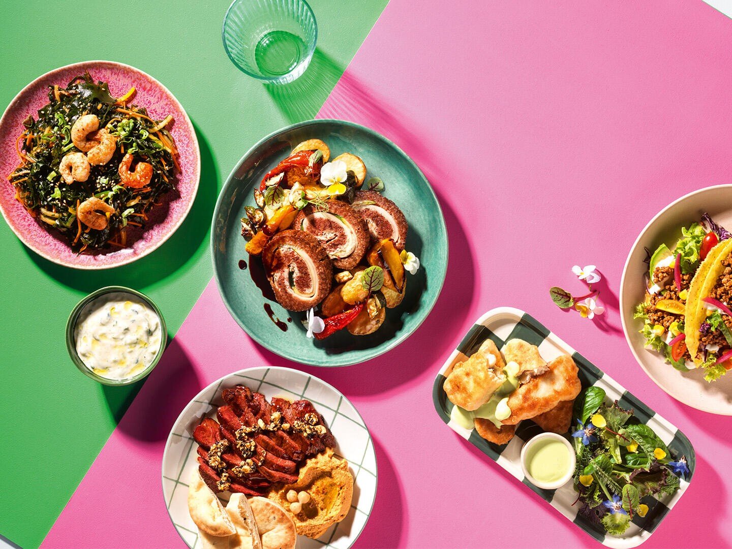 Auf einem farbenfrohen Tisch stehen verschiedene Gerichte, darunter Salat mit Garnelen, ein Teller mit gerolltem Fleisch und Gemüse, Steakscheiben mit Hummus und Brot, eine kleine Schale mit Dip und ein Tablett mit gebratenen Snacks und Salat auf leuchtend rosa und grünen Hintergründen.