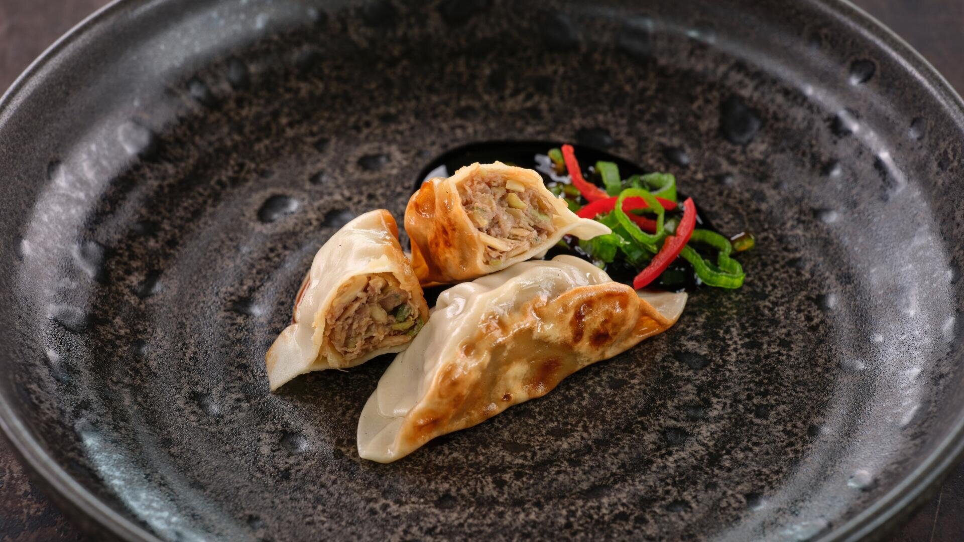 Drei in der Pfanne gebratene Knödel werden auf einem dunkel strukturierten Teller angerichtet, wobei ein Knödel in der Mitte durchgeschnitten ist, um die Füllung zu zeigen. Dünn geschnittenes rotes und grünes Gemüse garniert die Beilage.