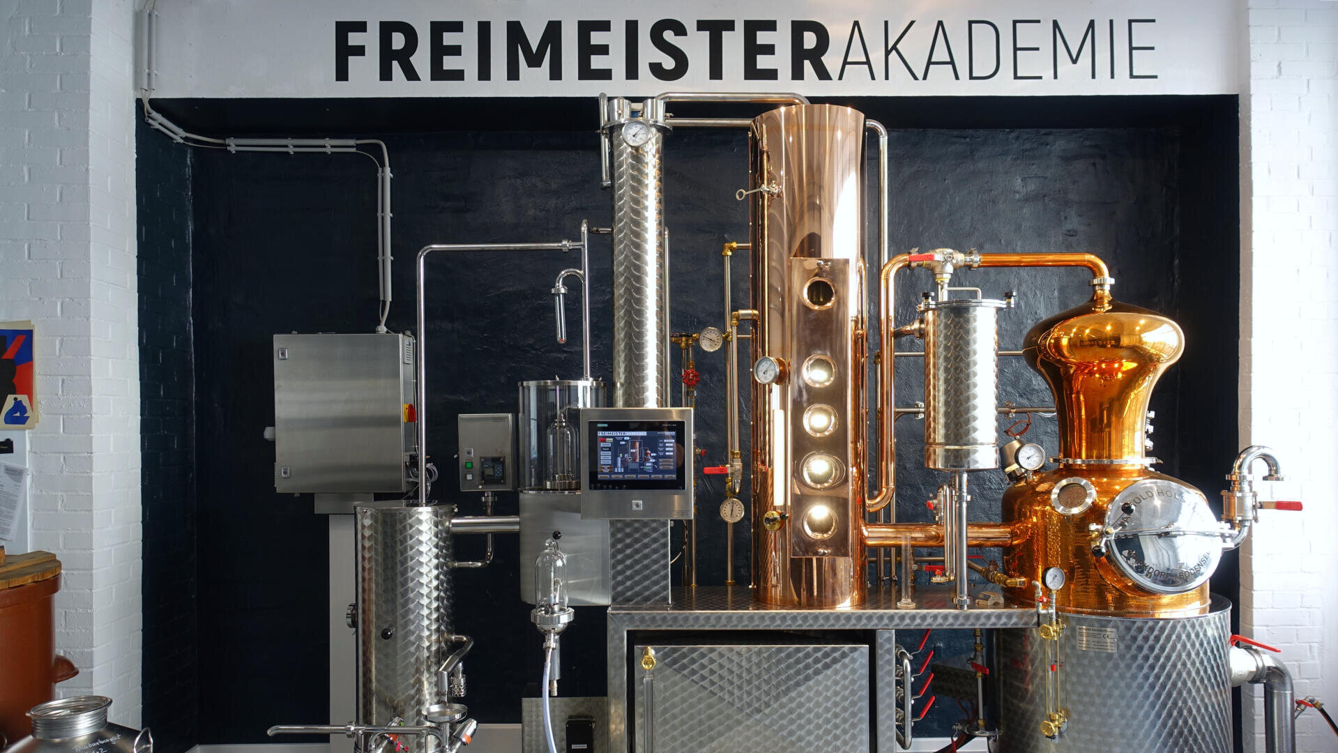 Un appareil de distillation moderne avec des composants en métal et en cuivre se trouve sous un panneau indiquant FREIMEISTERAKADEMIE sur un mur noir dans un environnement industriel lumineux.