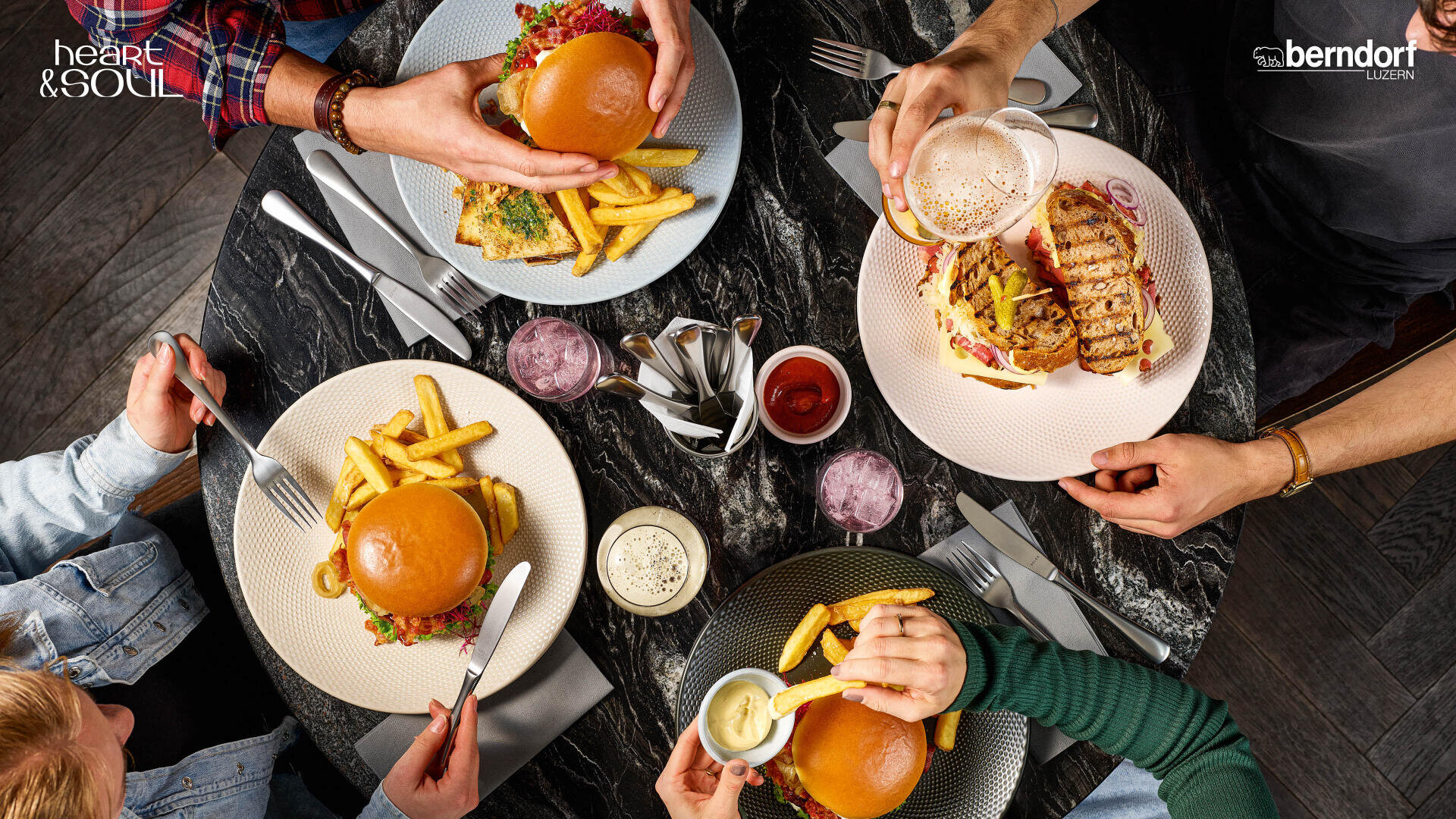 Vier Personen sitzen um einen dunklen Marmortisch und essen Hamburger, Pommes frites und ein Sandwich, dazu gibt es Getränke. Die Teller, das Besteck und die Soßen sind ordentlich sortiert und die Hände strecken sich nach dem Essen aus. Die Logos befinden sich in den oberen Ecken des Bildes.