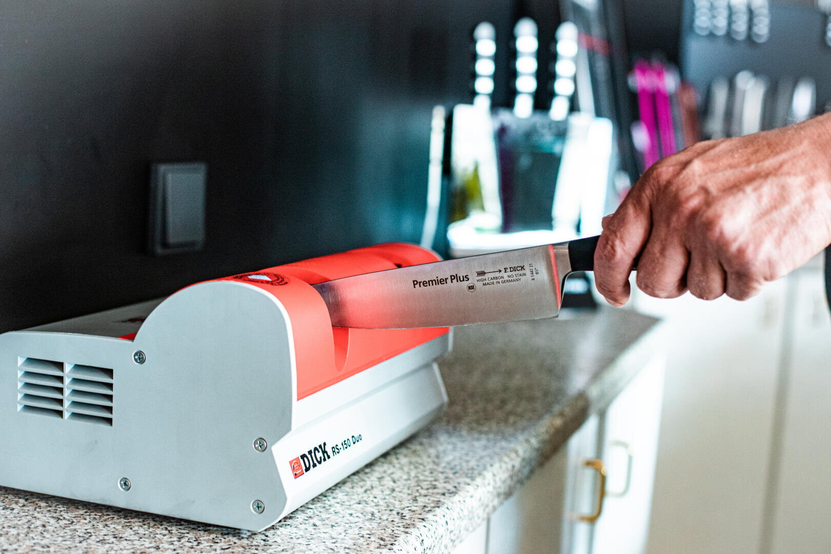 Eine Person schärft ein großes Küchenmesser mit einem elektrischen Messerschärfgerät auf einer Arbeitsplatte, wobei verschiedene Utensilien im Hintergrund verschwimmen.