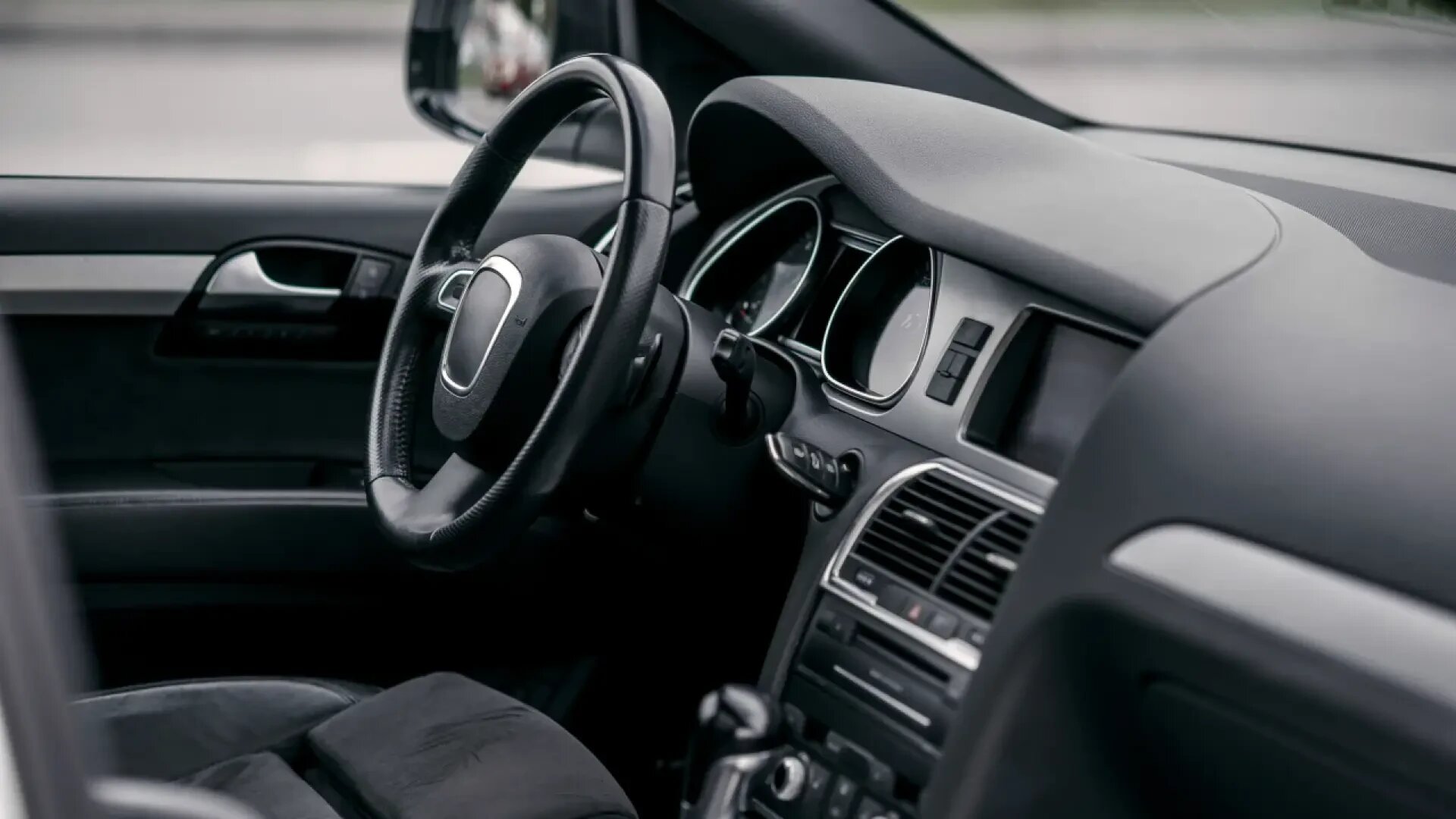 Nahaufnahme des Innenraums eines modernen Fahrzeugs, die das Lenkrad, das Armaturenbrett, die Mittelkonsole und einen Teil des Fahrersitzes in einem sauberen, schwarz-grauen Farbschema zeigt.