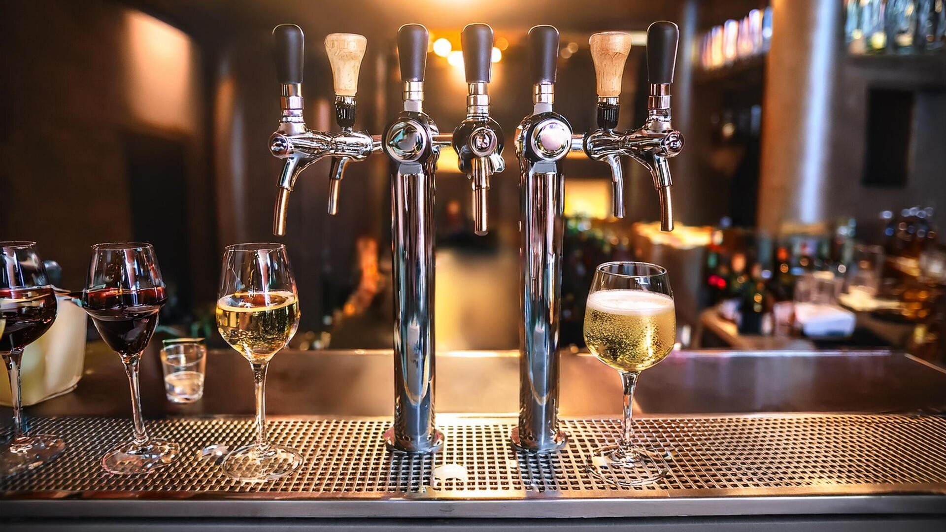 Nahaufnahme von Bierzapfhähnen auf einem Bartresen mit Gläsern Rot- und Weißwein und einem Glas Bier, mit warmer Beleuchtung und unscharfen Flaschen im Hintergrund.